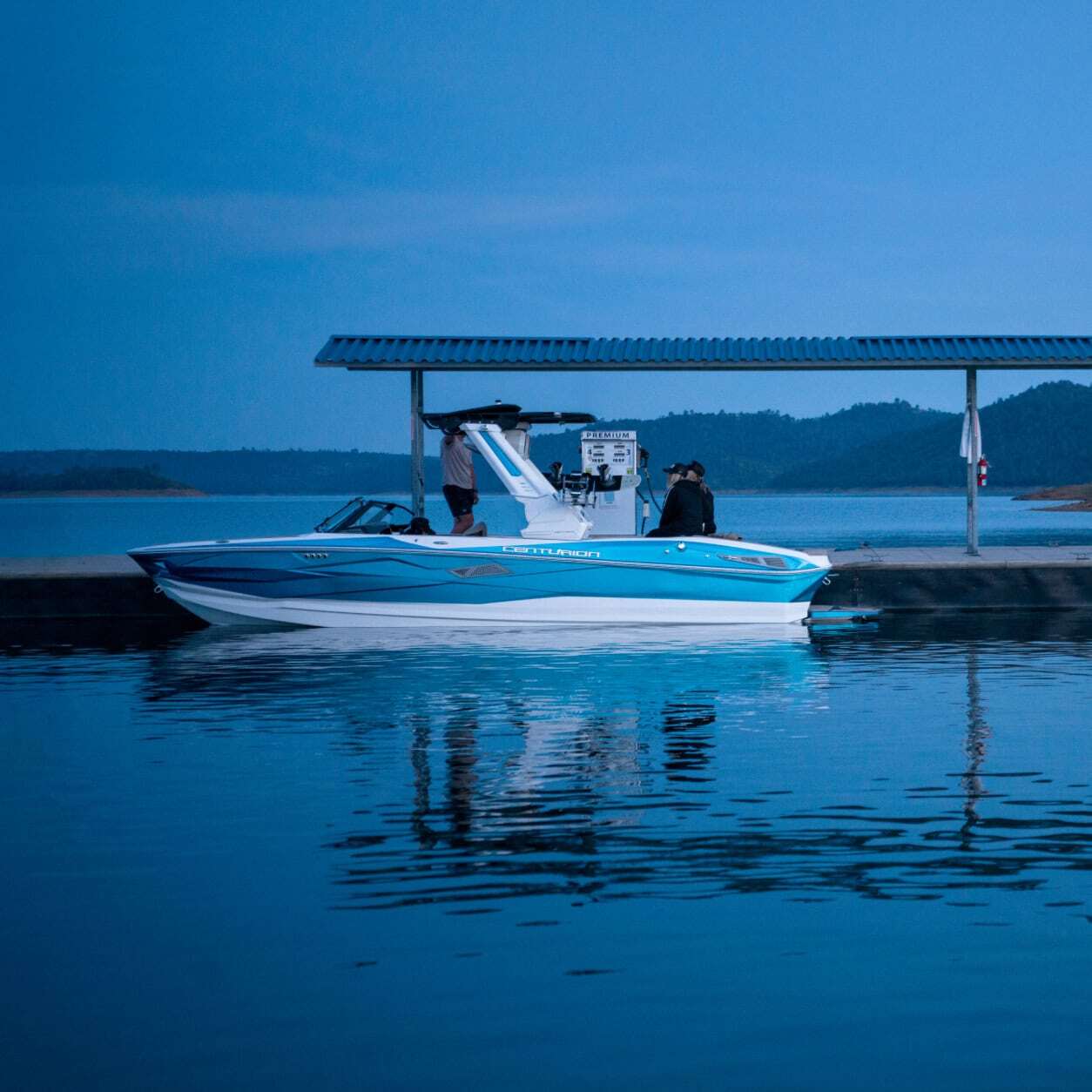 A blue and white speedboat is docked beside a covered fuel station on a calm lake at dusk, with hills visible in the background.
