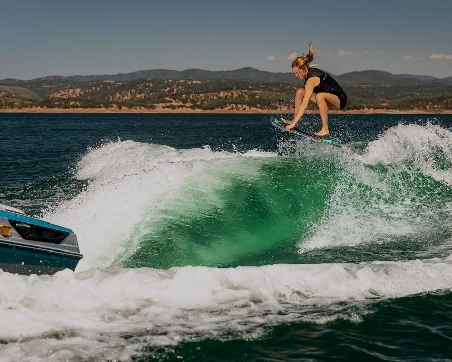 A person in a wetsuit is wakesurfing, catching air above the water behind a boat equipped with Centurion Ballast on a lake with hills in the background.