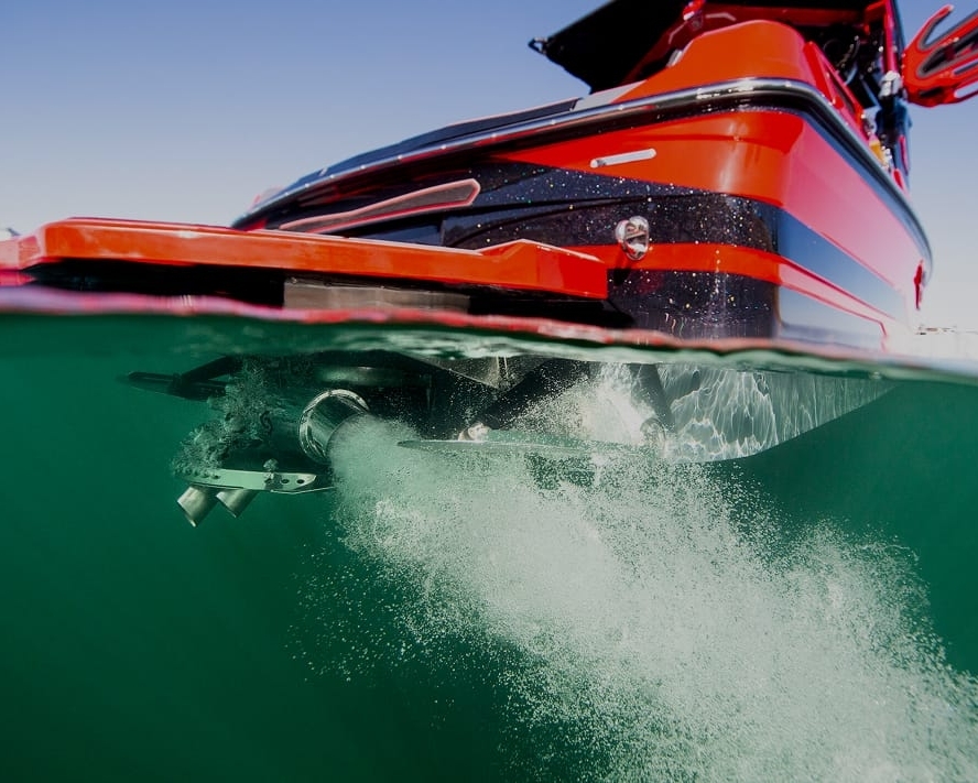 A red boat with a Centurion Opti-V Hull is partially submerged in water, bubbles trailing from its propeller, as seen from a split above-and-below water perspective.