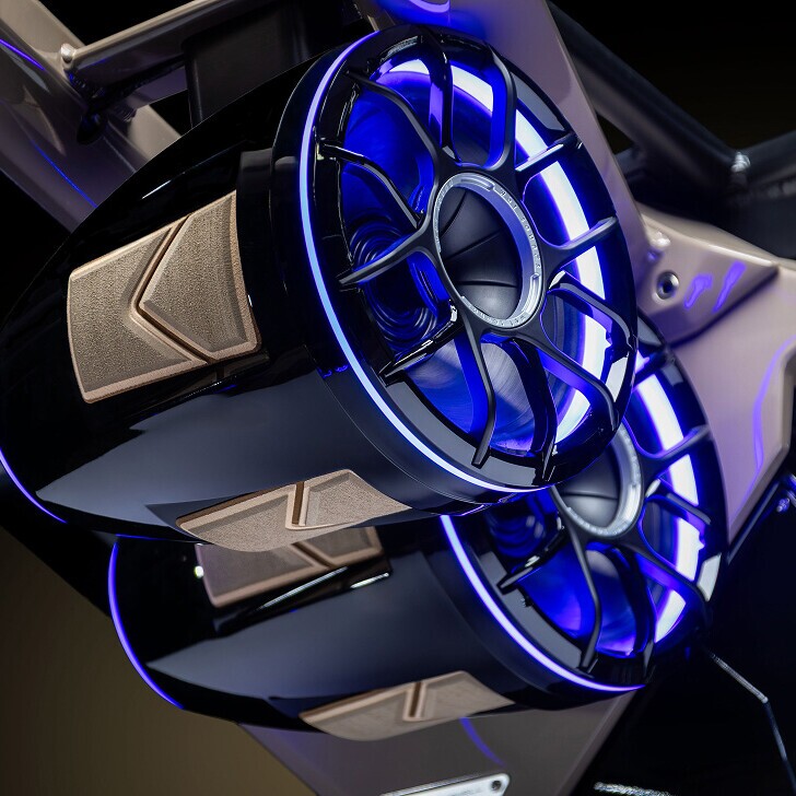 Close-up of two futuristic, blue LED-lit speakers with metallic accents, featuring a geometric design and positioned at an angle against a dark background.