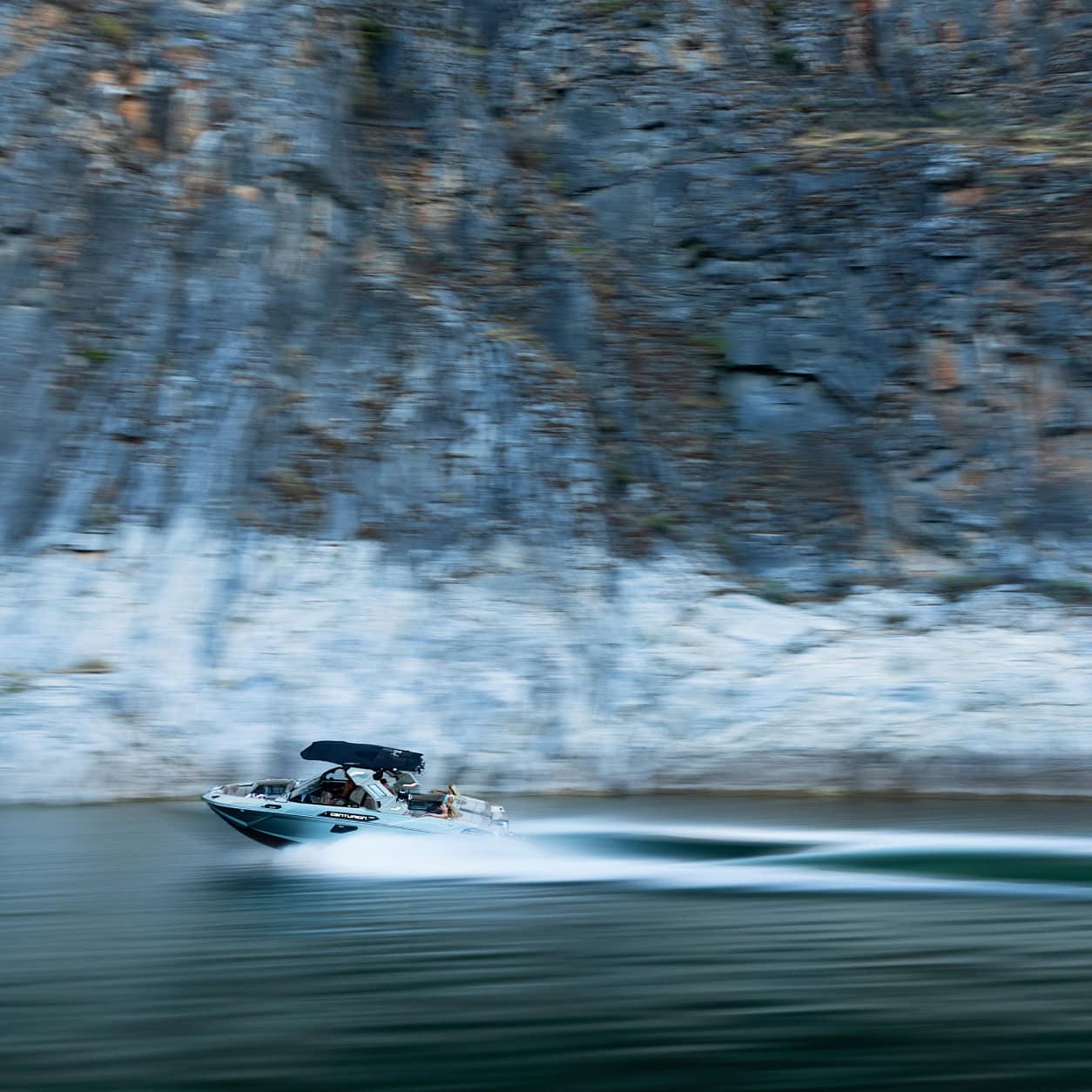 A speedboat moves quickly across a calm body of water, creating a blurred motion effect, with tall rocky cliffs in the background.