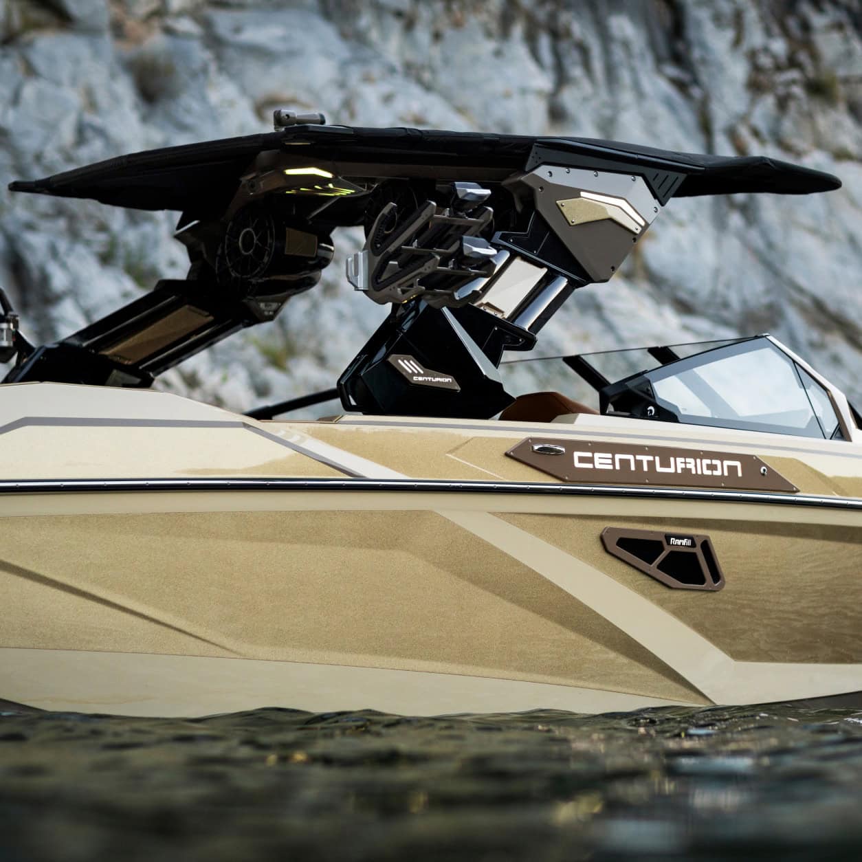 A tan Centurion boat floats on the water near rocky cliffs, showing a close-up of its side, windshield, and wakeboard tower.