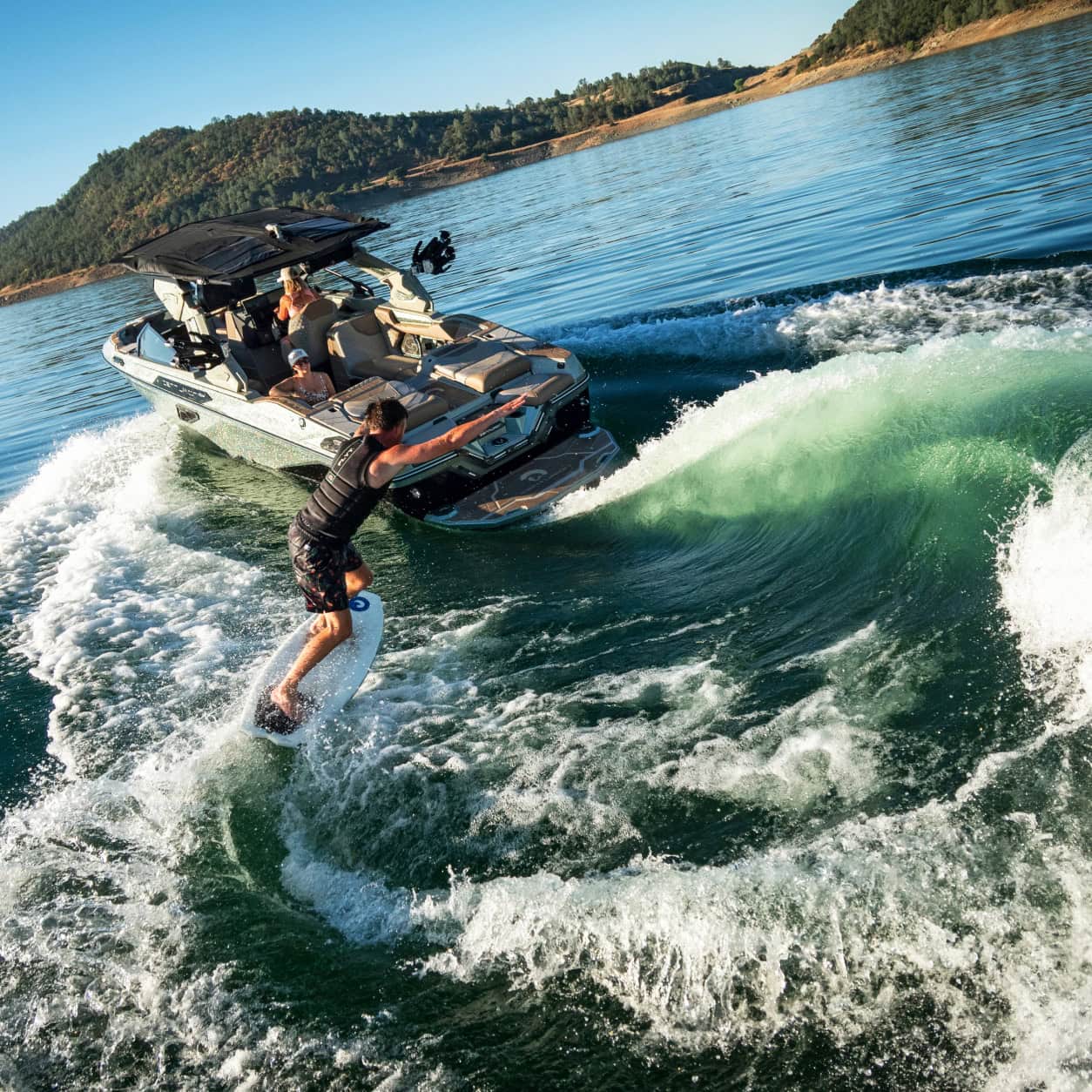 A person wake surfs on a wave behind a motorboat on a lake, surrounded by forested hills under a clear blue sky.