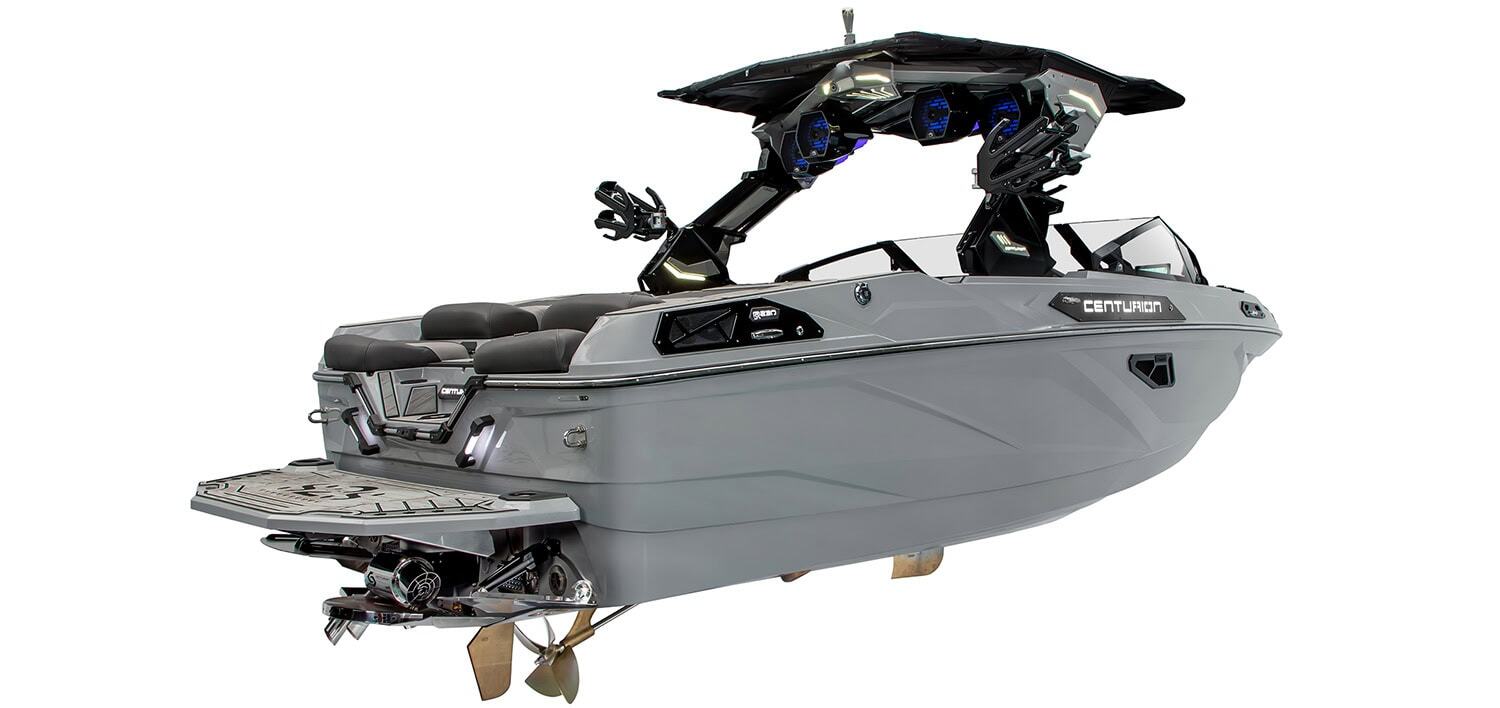 Gray wakeboard boat with black accents, rear swim platform, and seating; features tower with speakers and racks for boards, viewed from rear side angle.