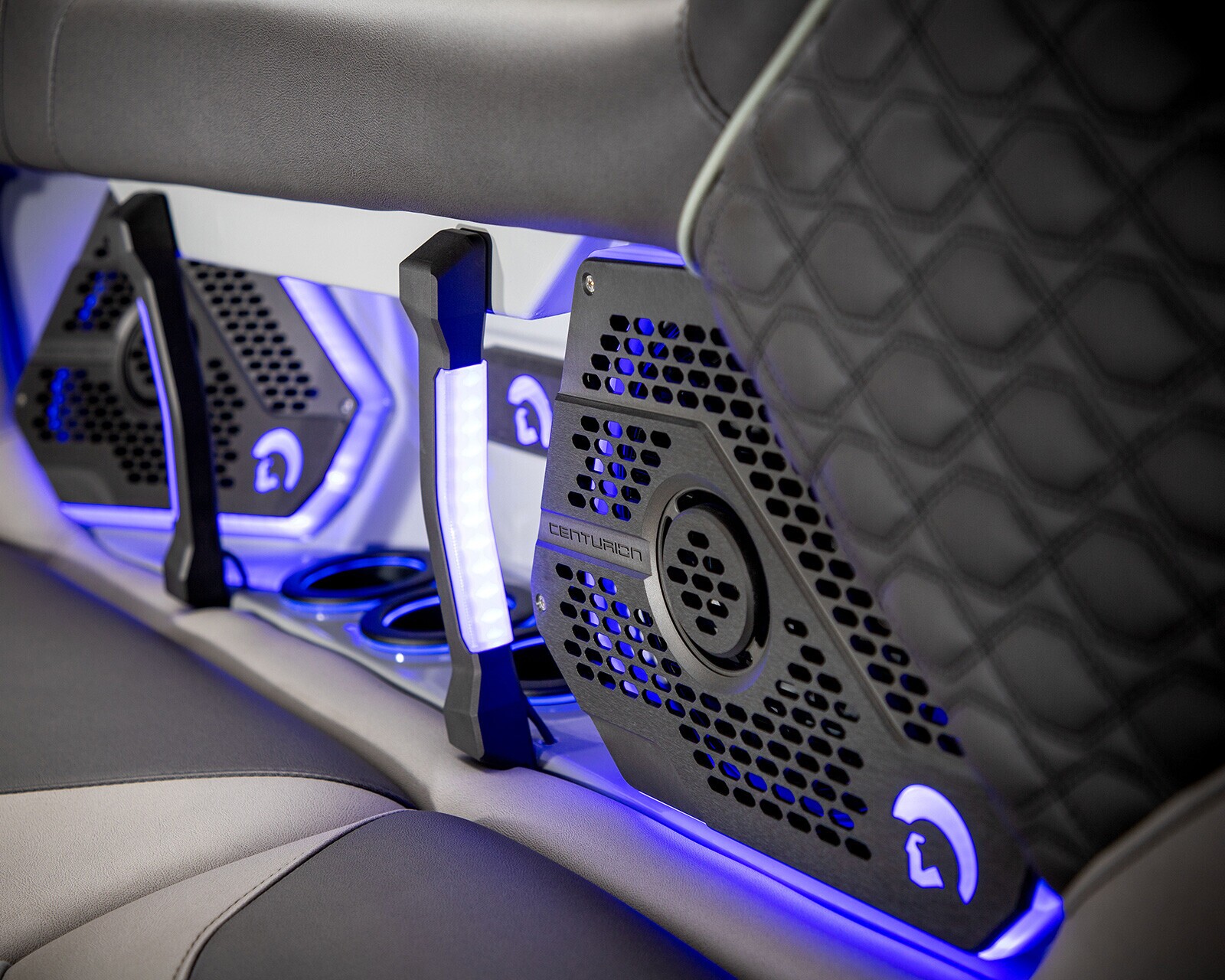 Close-up of a car or boat interior showing black and gray quilted upholstery, illuminated cup holders, and speakers with blue LED lighting.