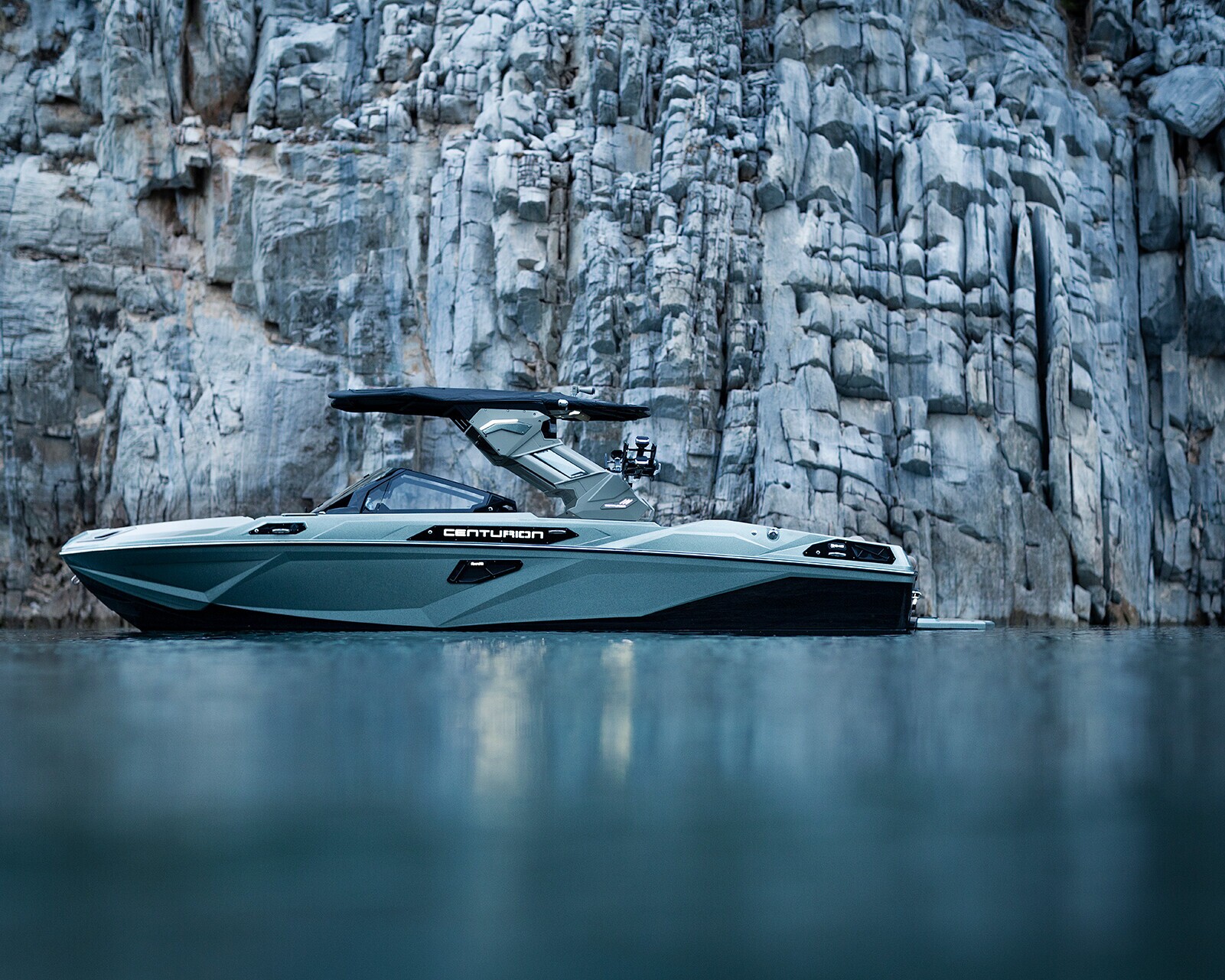A modern Centurion boat floats on calm water near a steep, rocky cliff.
