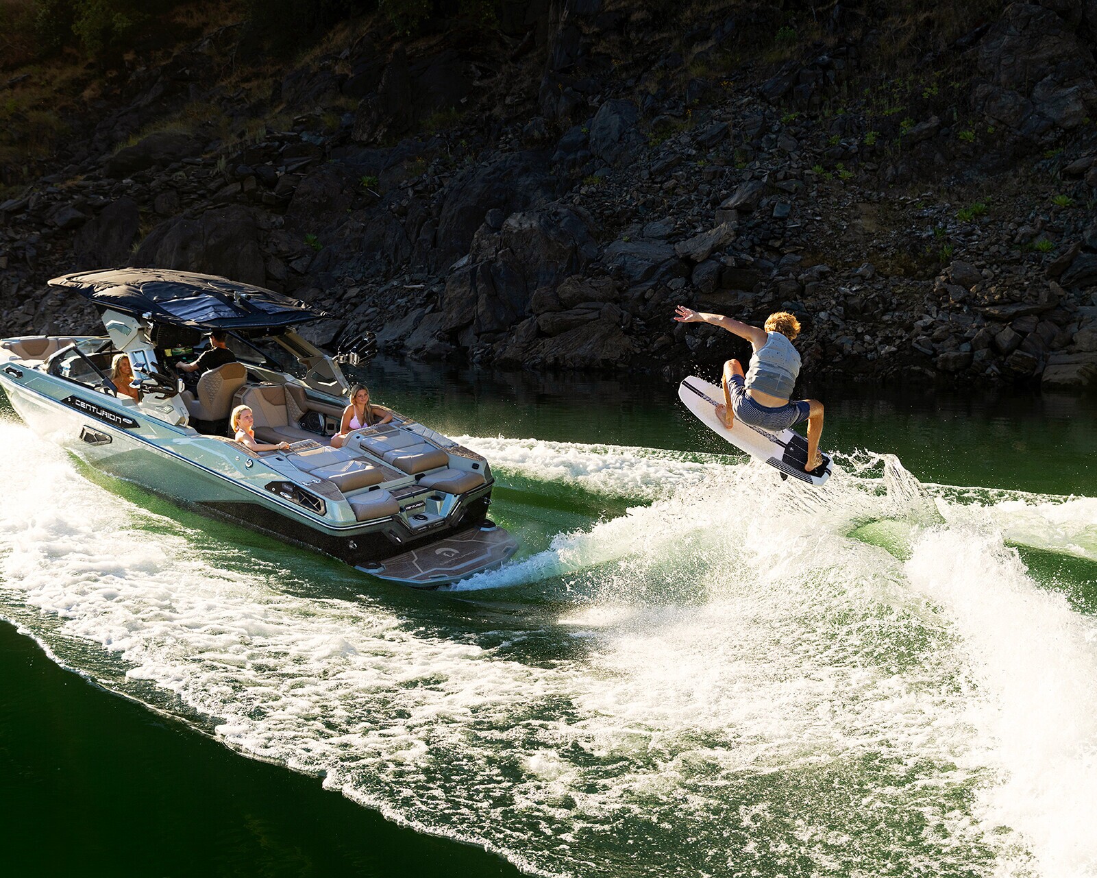 A person rides a wakeboard behind a motorboat on a lake, while several people watch from inside the boat. Rocky shoreline is visible in the background.