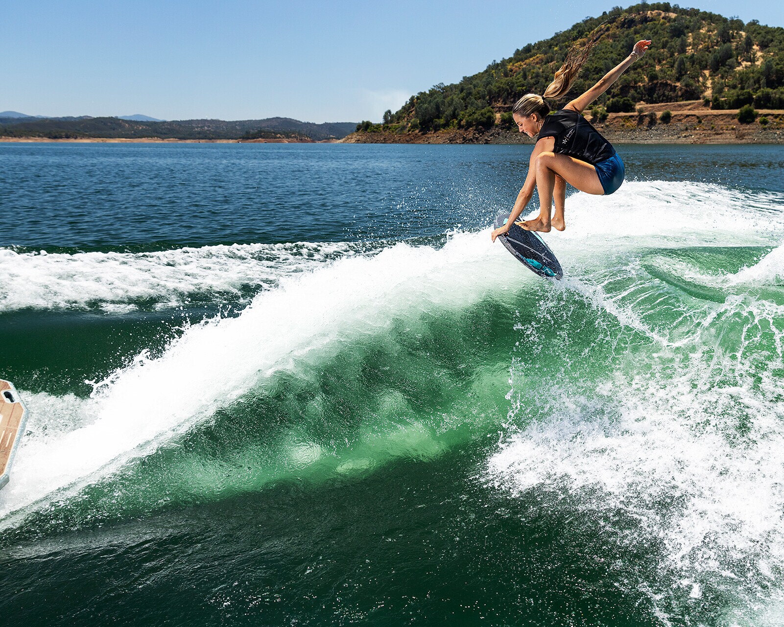 A person wakesurfs on a lake near a boat, jumping off a wave with hills and blue sky visible in the background.