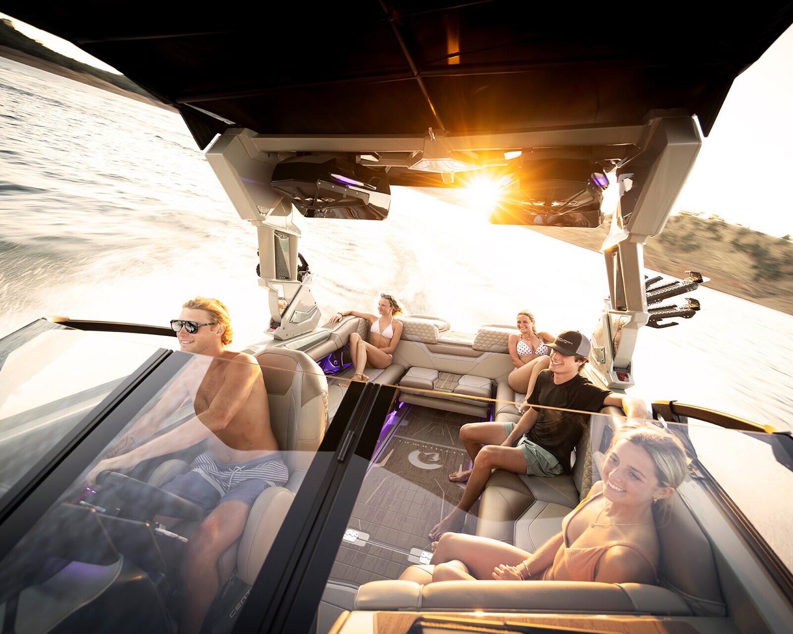 Five people ride on a speedboat at sunset, with water splashing behind them. Some are seated, others are relaxing, and the driver is steering. The scene is bright and energetic.