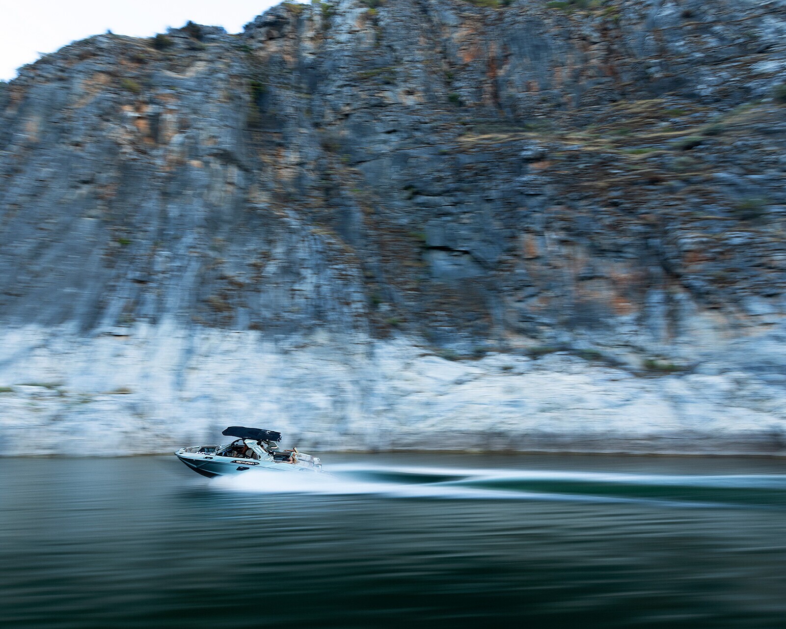 A small motorboat speeds across a calm body of water, with blurred motion, near steep rocky cliffs.
