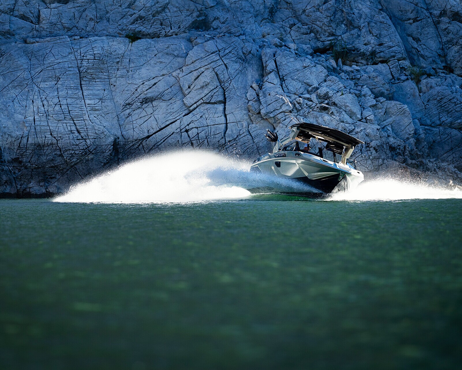 A speedboat moves quickly across a green lake, creating a large spray of water, with rocky cliffs visible in the background.