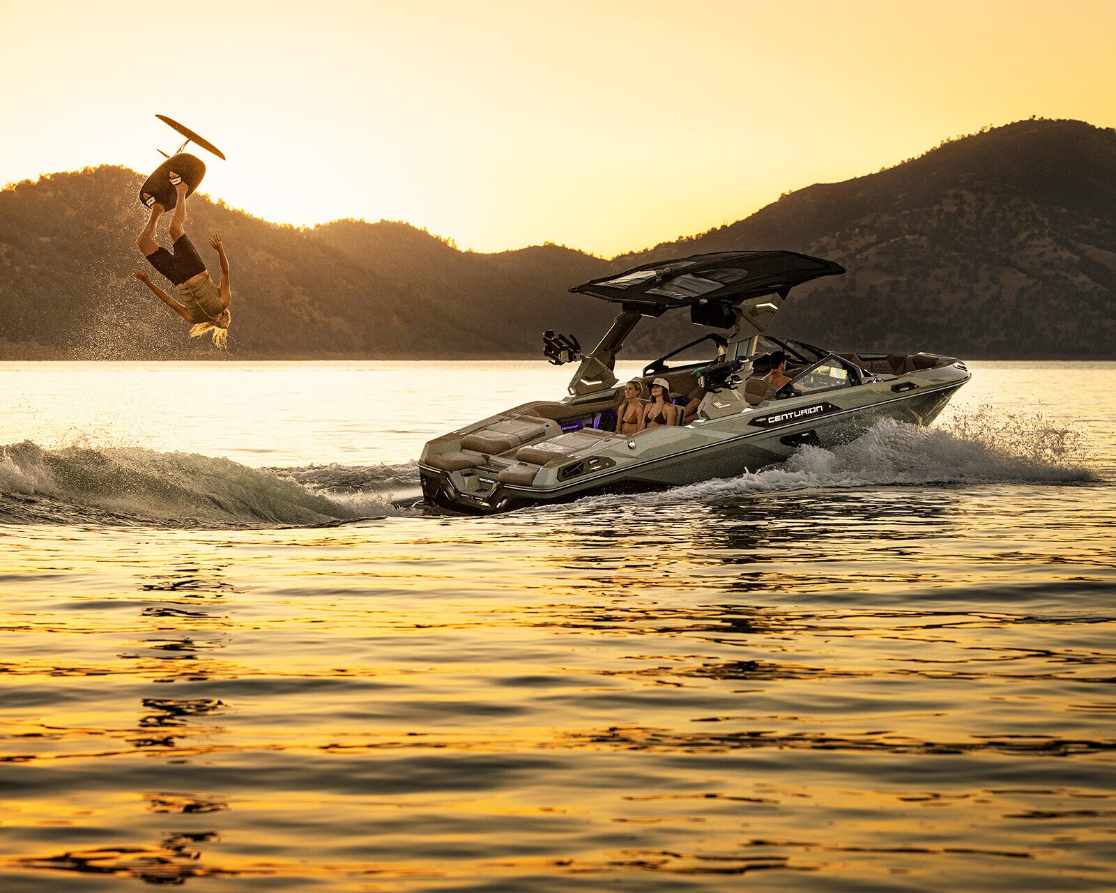 A person wakesurfs in mid-air behind a speedboat on a lake at sunset, with mountains visible in the background.