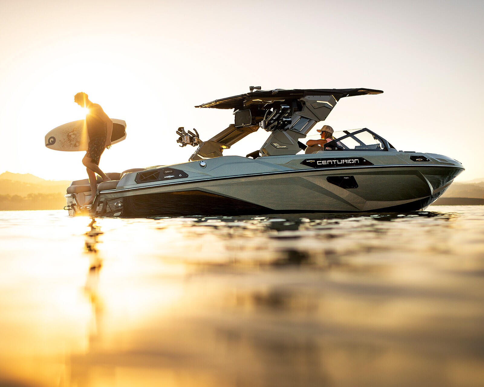 A person stands on a boat holding a surfboard at sunset, with another person seated inside the boat on calm water.