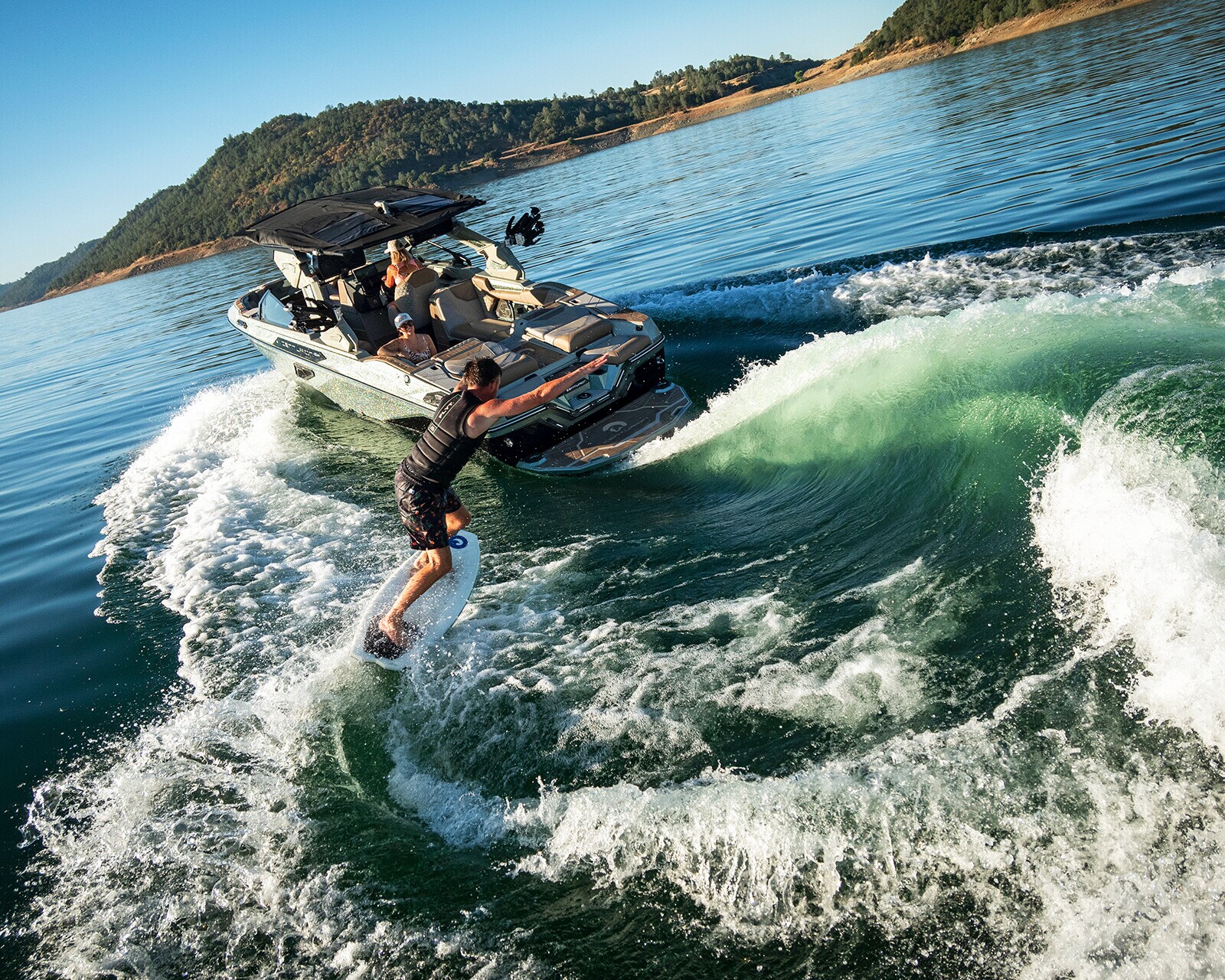 A person wake surfs behind a motorboat on a lake, with hills and clear sky in the background.