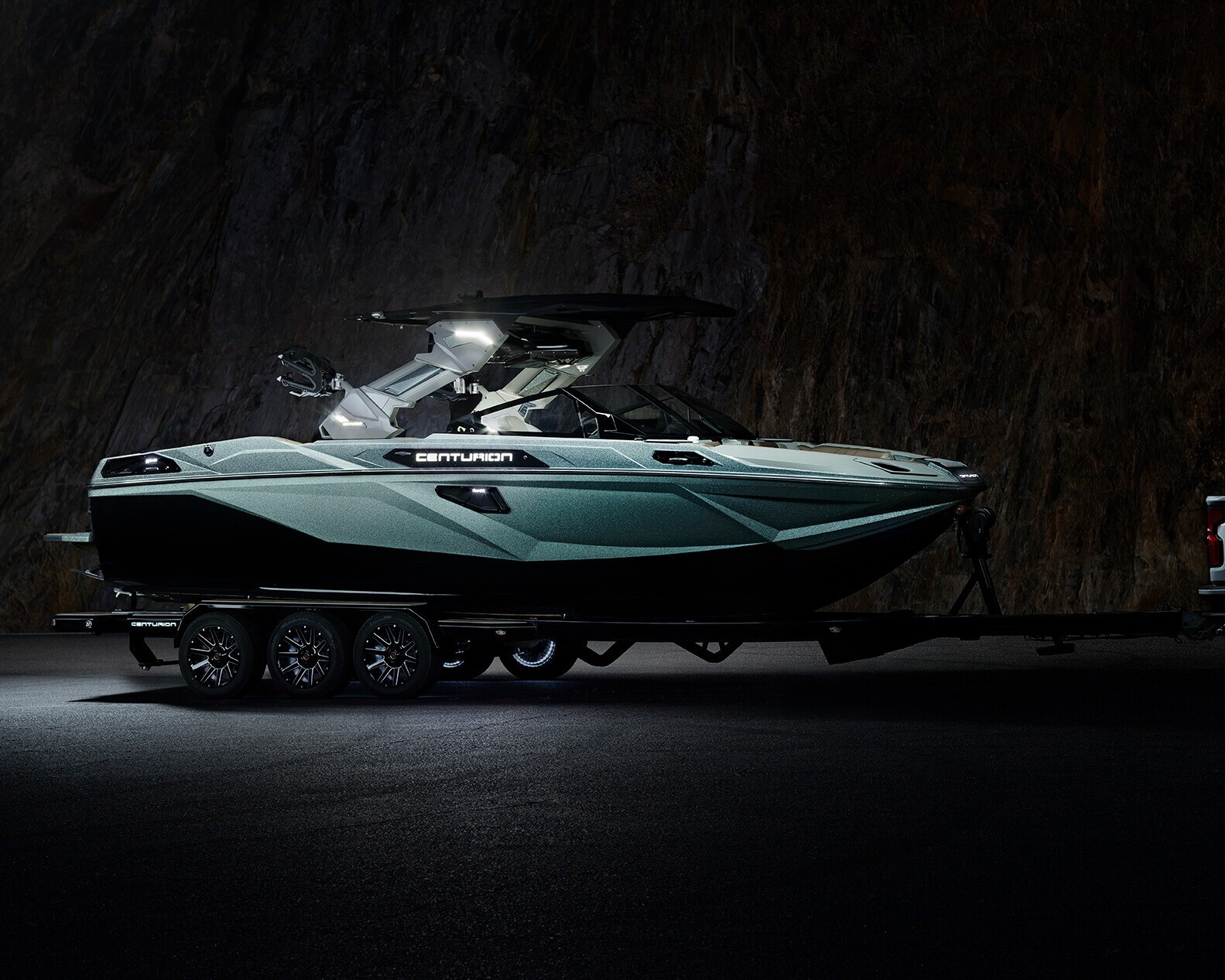 A dark green Camryon boat sits on a triple-axle trailer, hitched to a truck, illuminated against a rocky background at night.