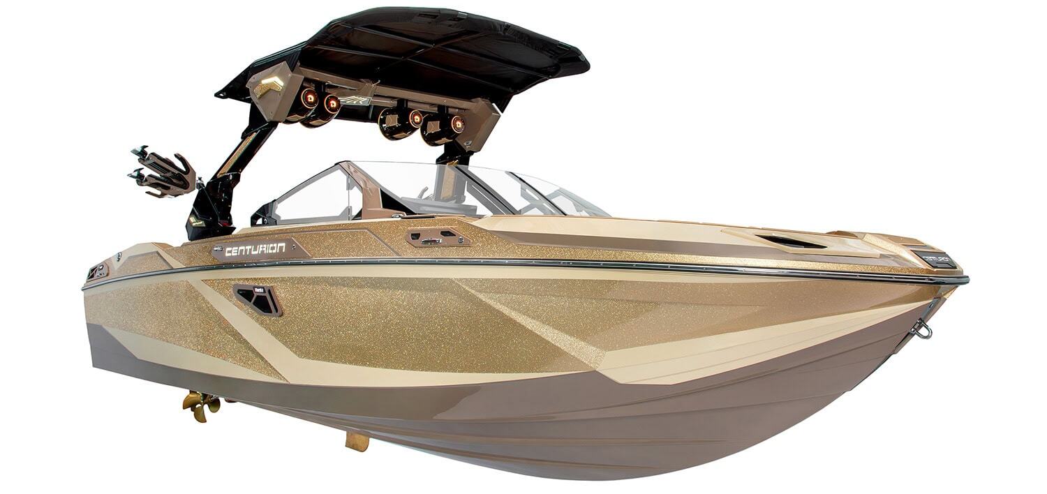 Gold and tan Centurion wakeboard boat with a black tower, angled bow, and metallic finish, viewed from the front side against a white background.