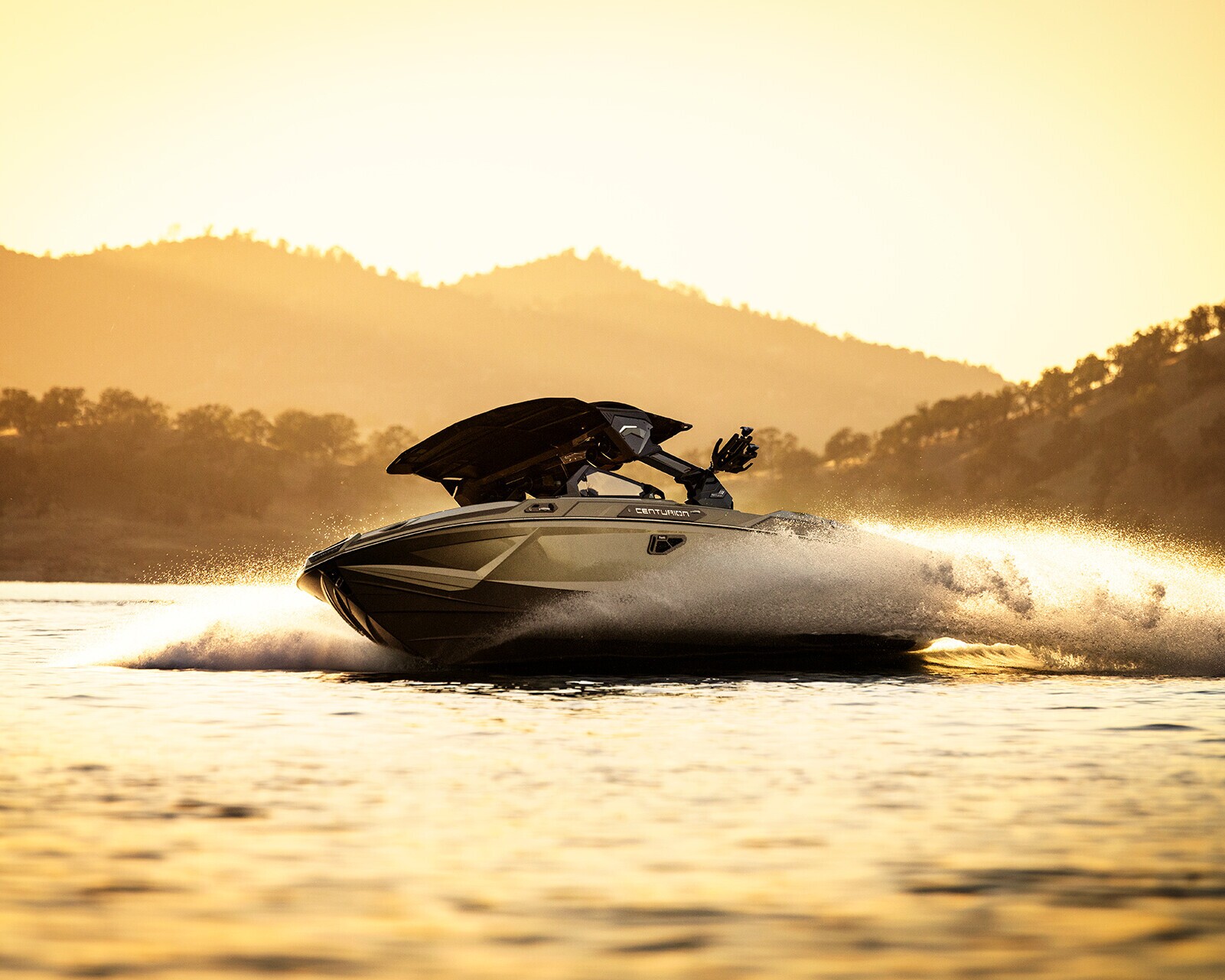 A speedboat moves quickly across a lake at sunset, creating a spray of water behind it, with hills and trees visible in the background.