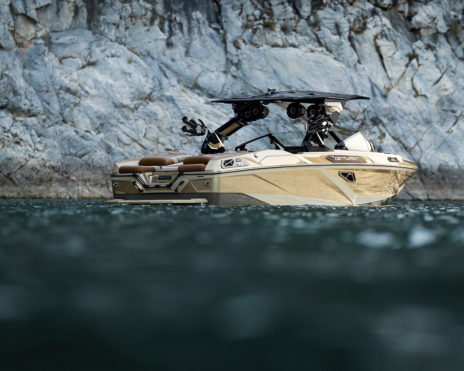 A tan and black wakeboard boat floats on green water near a rocky cliffside.
