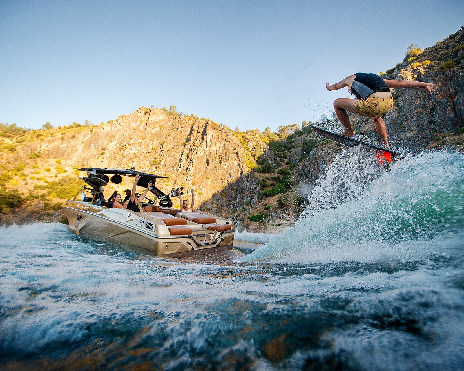 A person on a wakeboard jumps over a wave behind a speedboat with several passengers, set against rocky hills and blue sky.