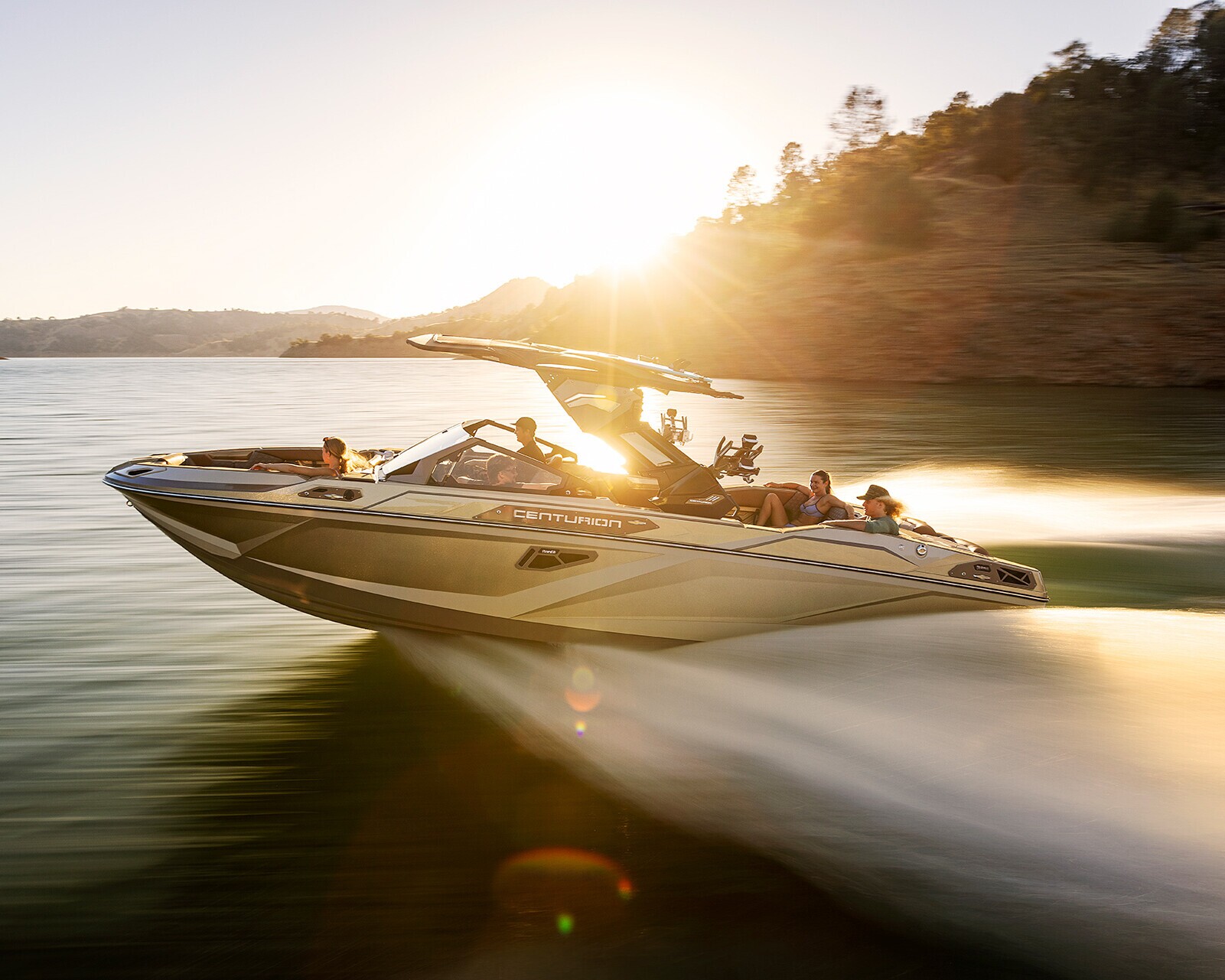 A speedboat with several passengers moves quickly across a lake at sunset, creating a wake, with a rocky shoreline and trees in the background.