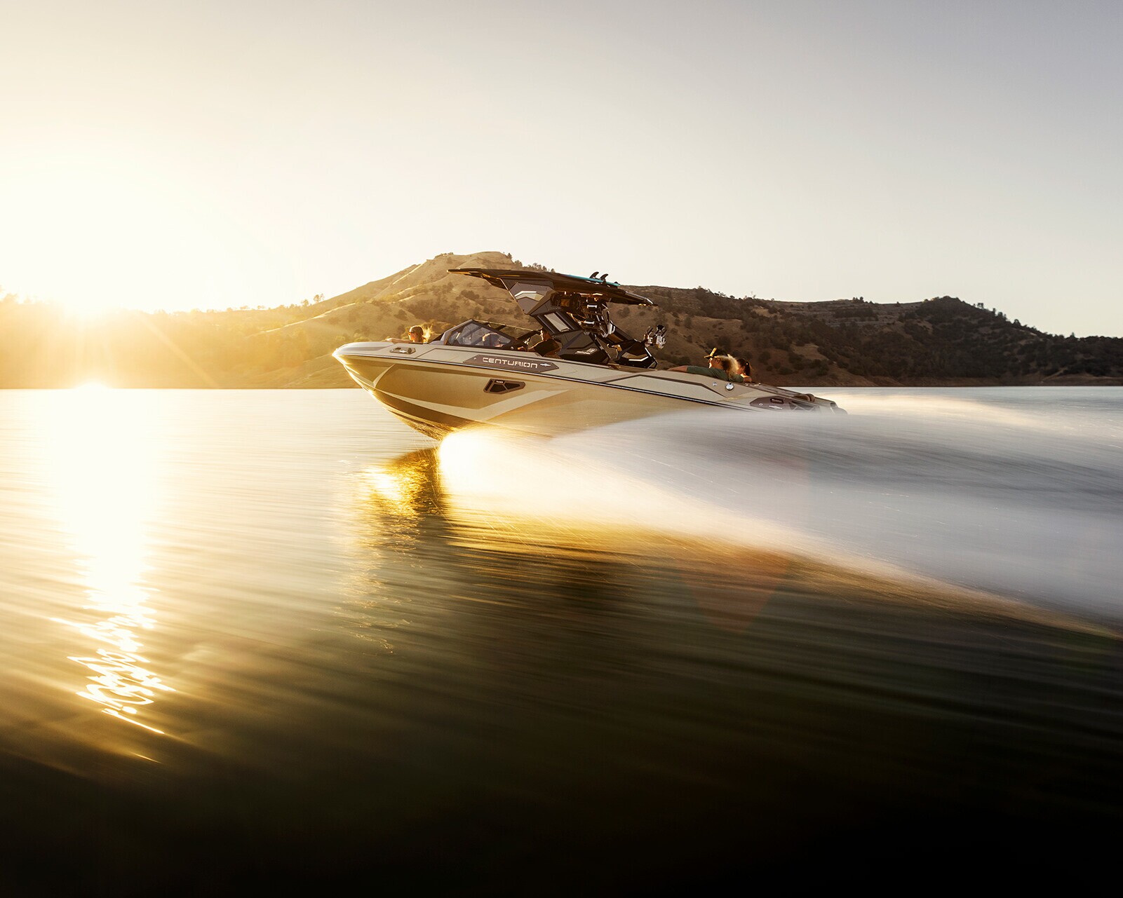 A speedboat moves quickly across a calm lake at sunset, creating a wake behind it with hills and the sun in the background.