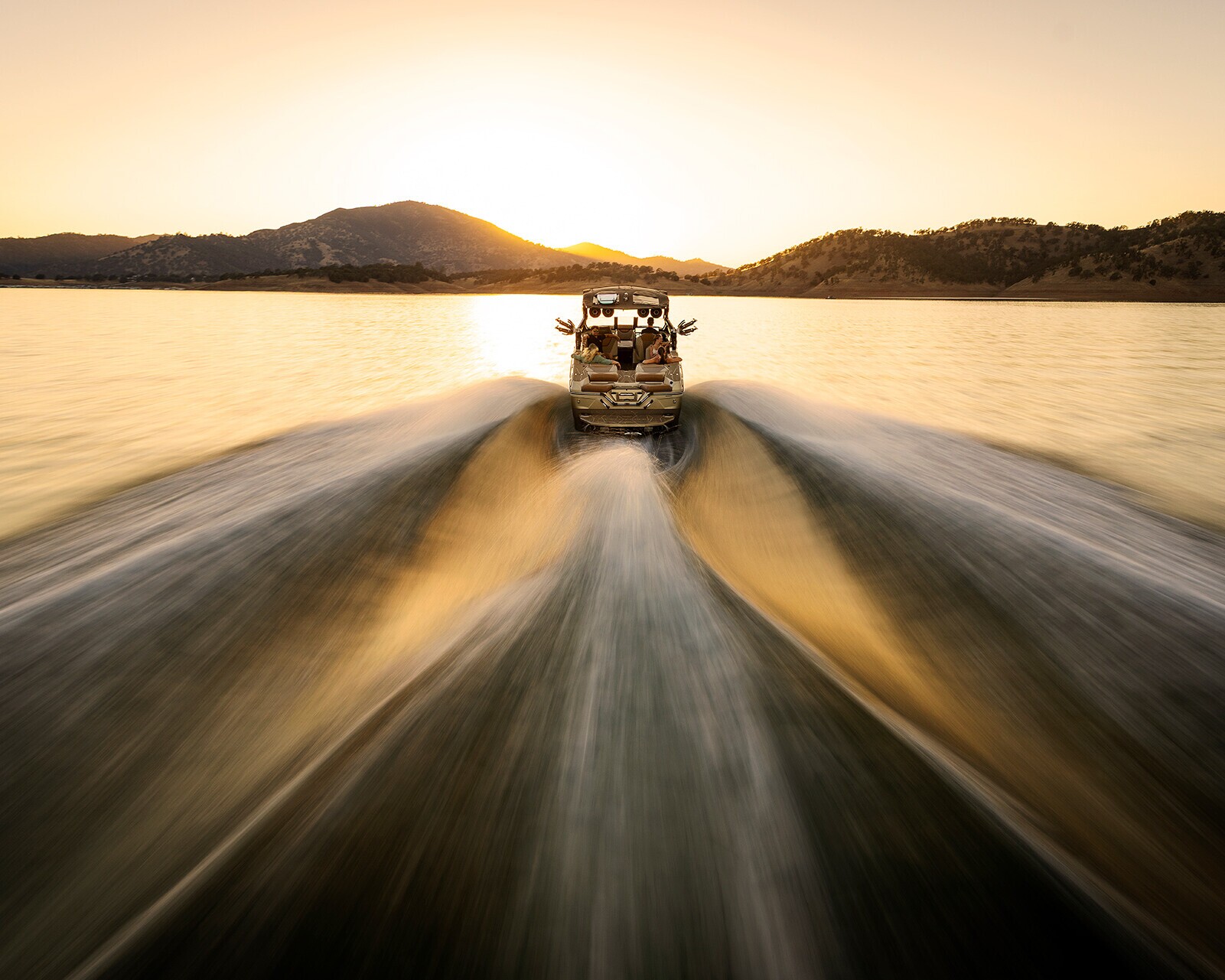 A speedboat moves across a lake at sunset, leaving trails in the water, with hills in the background.