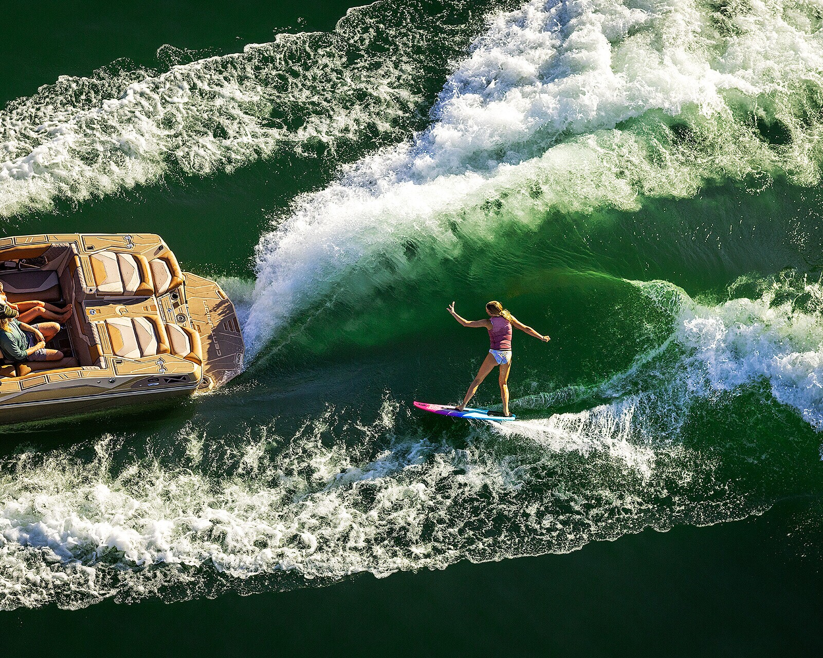 A person wake surfs behind a tan motorboat on green water, balancing on a surfboard as the boat creates large waves.