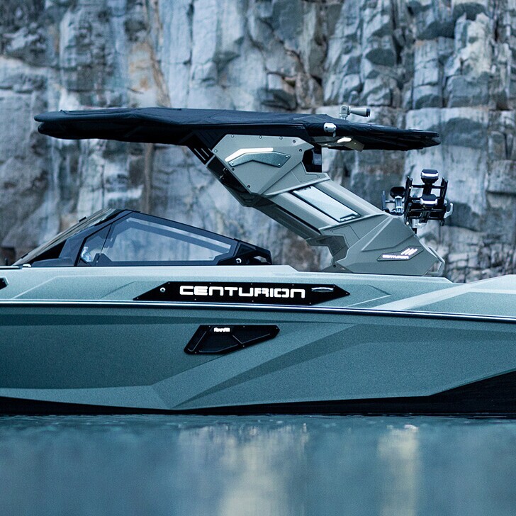 Side view of a Centurion speedboat with a black canopy, floating on calm water near a rocky cliff.