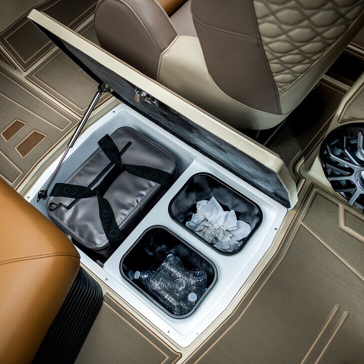 Opened car floor compartment showing storage bins containing a bag, crumpled tissues, and water bottles. Brown and beige car interior visible.