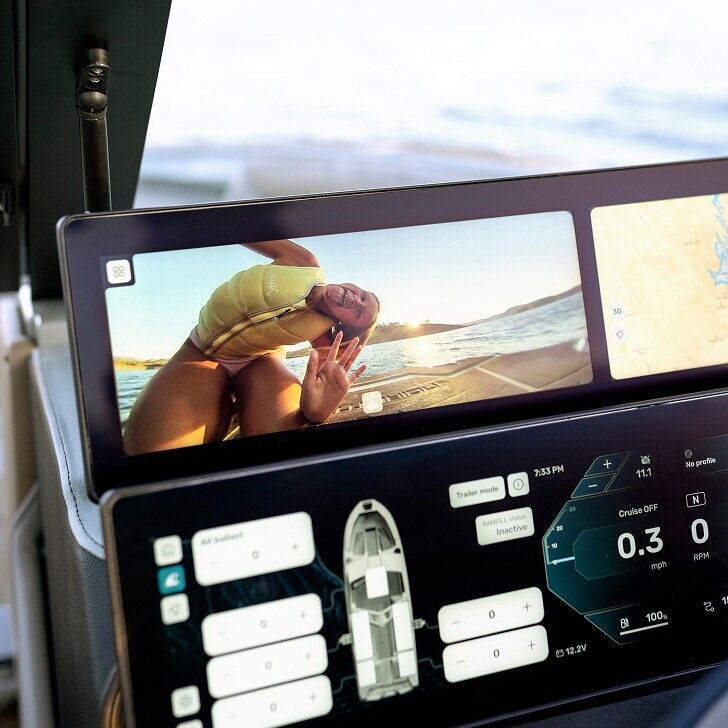 A boat dashboard display shows a rear camera view of a person in a yellow life jacket making a peace sign, with digital navigation and boat status information below.
