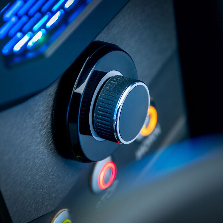 Close-up of a black rotary control knob on a dashboard, surrounded by several illuminated buttons in various colors.