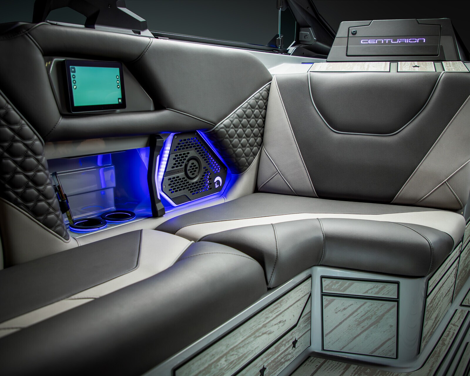 Modern boat interior featuring black and gray cushioned seating, cup holders, a control screen, a speaker with blue lighting, and