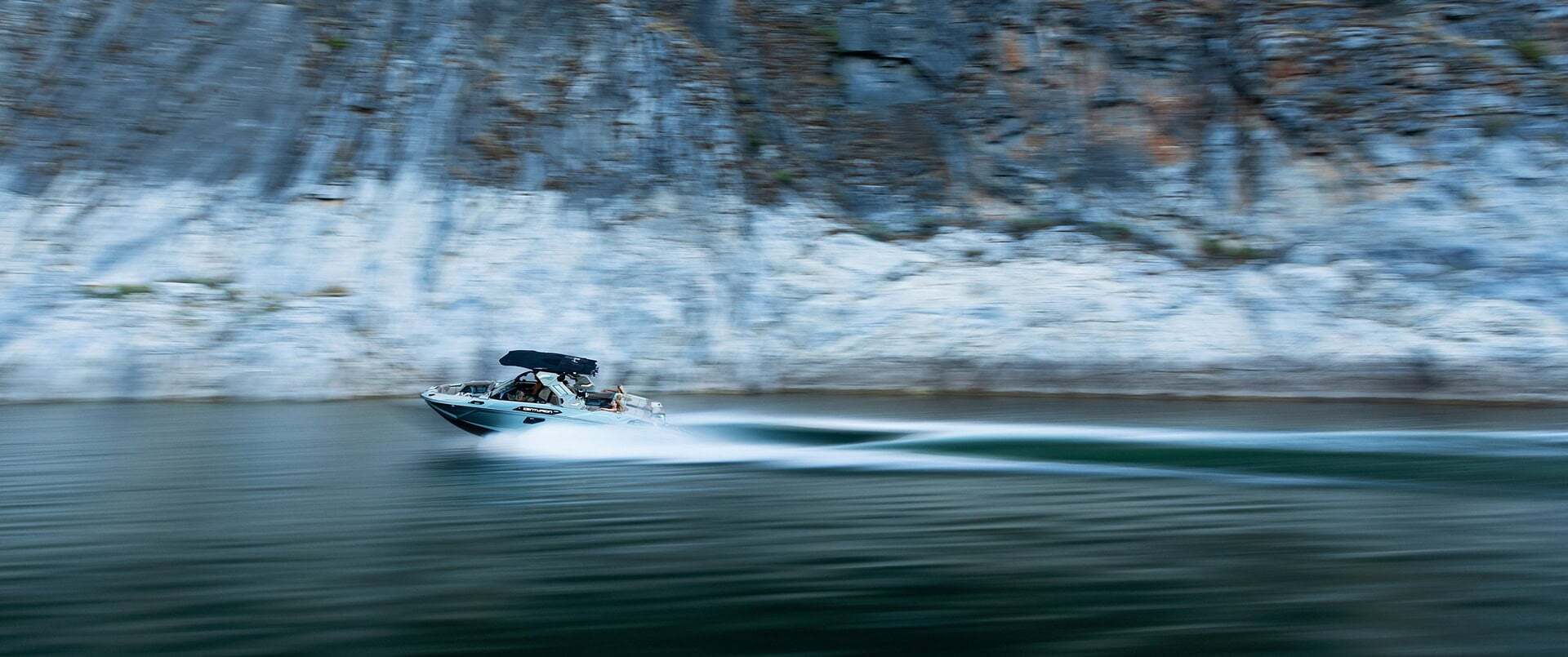 A small motorboat speeds across a calm body of water, leaving a wake behind, with rocky cliffs in the background.