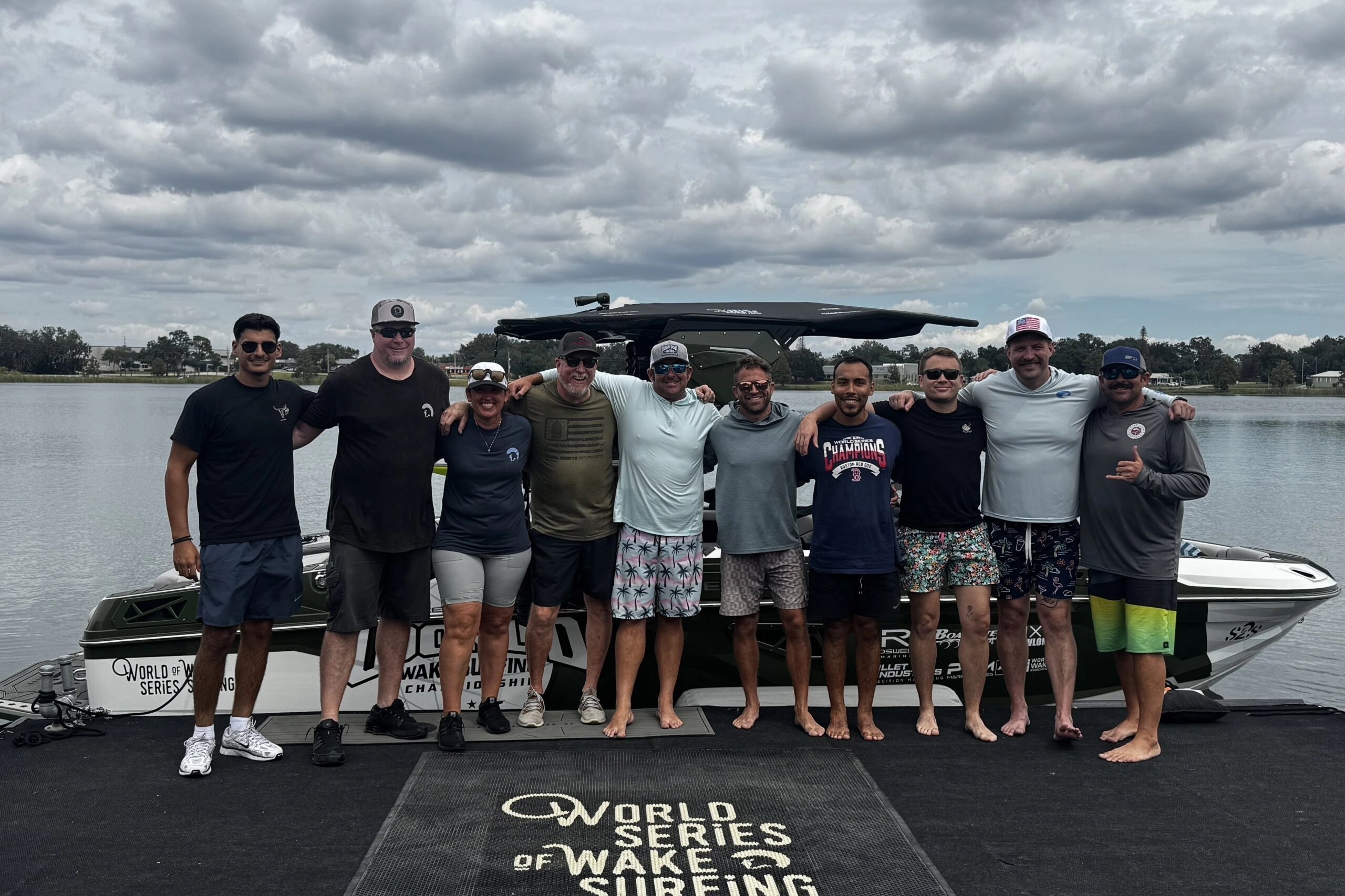 Ten people stand side by side on a dock in front of a boat, smiling, with a “World Series of Wake Surfing” logo on the dock surface and a lake in the background under a cloudy sky.