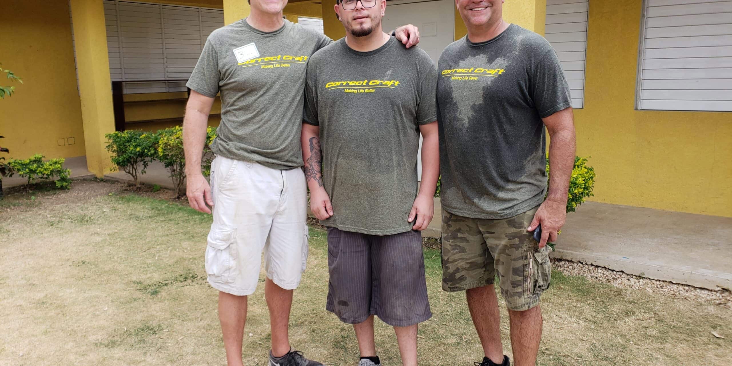 Three men wearing matching green Correct Craft t-shirts standing in front of a yellow building.
