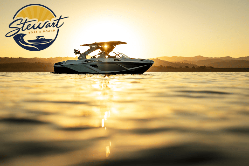 A Centurion wake boat floating on calm water at sunset, with the Stewart Boat & Board logo in the upper left corner.