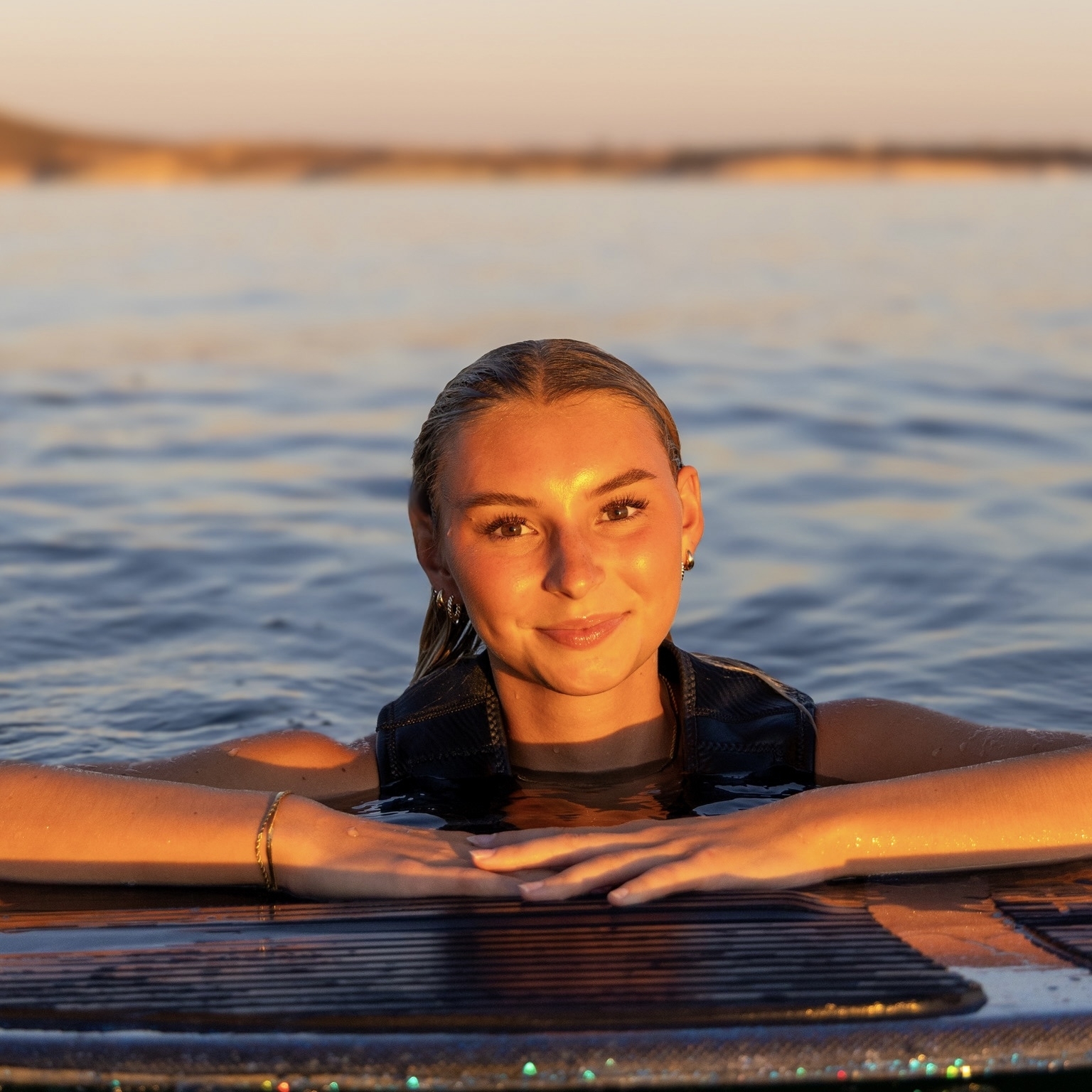 Ava Stewart, wearing a life vest, rests her arms on a surfboard in calm water at sunset, with golden light reflecting on her face.