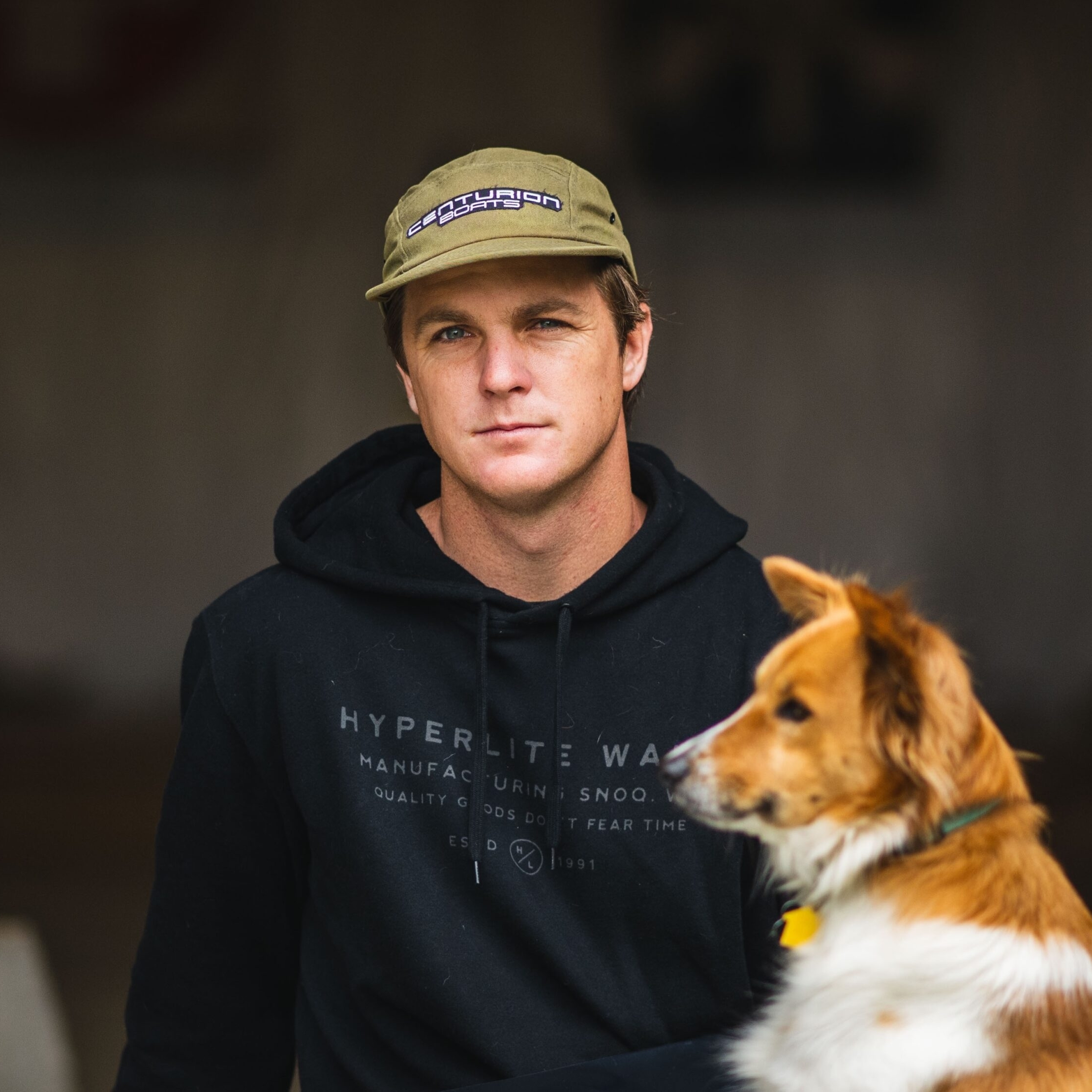 A man in a dark hoodie and cap sits indoors next to a brown and white dog, both facing forward.