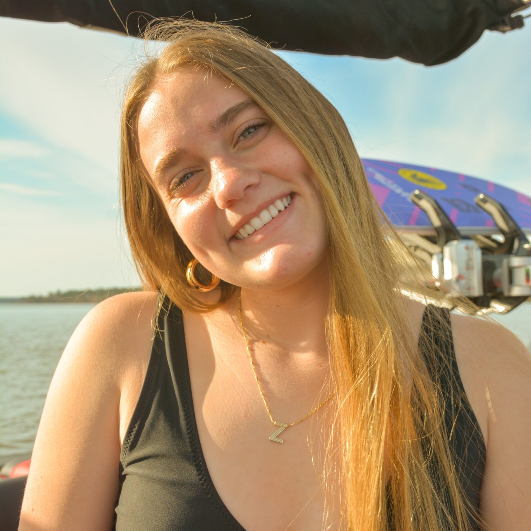 A young woman in a black bikini top smiles at the camera while sitting on a boat with water and blue sky in the background.