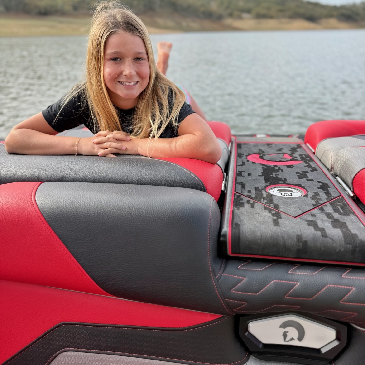 A young person with long blonde hair smiles while reclining on the red and gray seats of a boat on a lake, with hills and trees in the background.