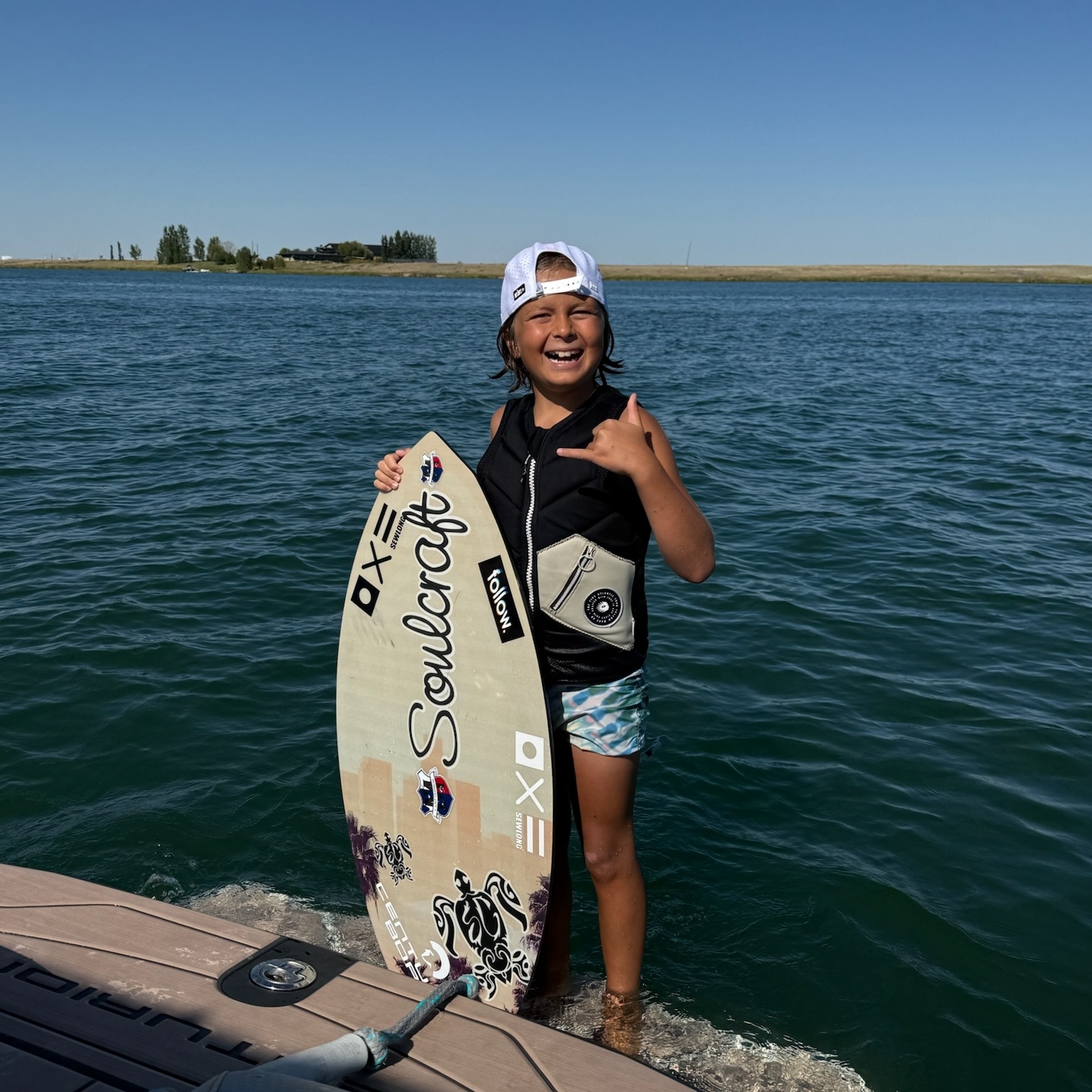 Young person in a life jacket and cap stands on a dock by the water, holding a wake surfboard and giving a thumbs up on a sunny day—ready for action just like Greyson Hantke.