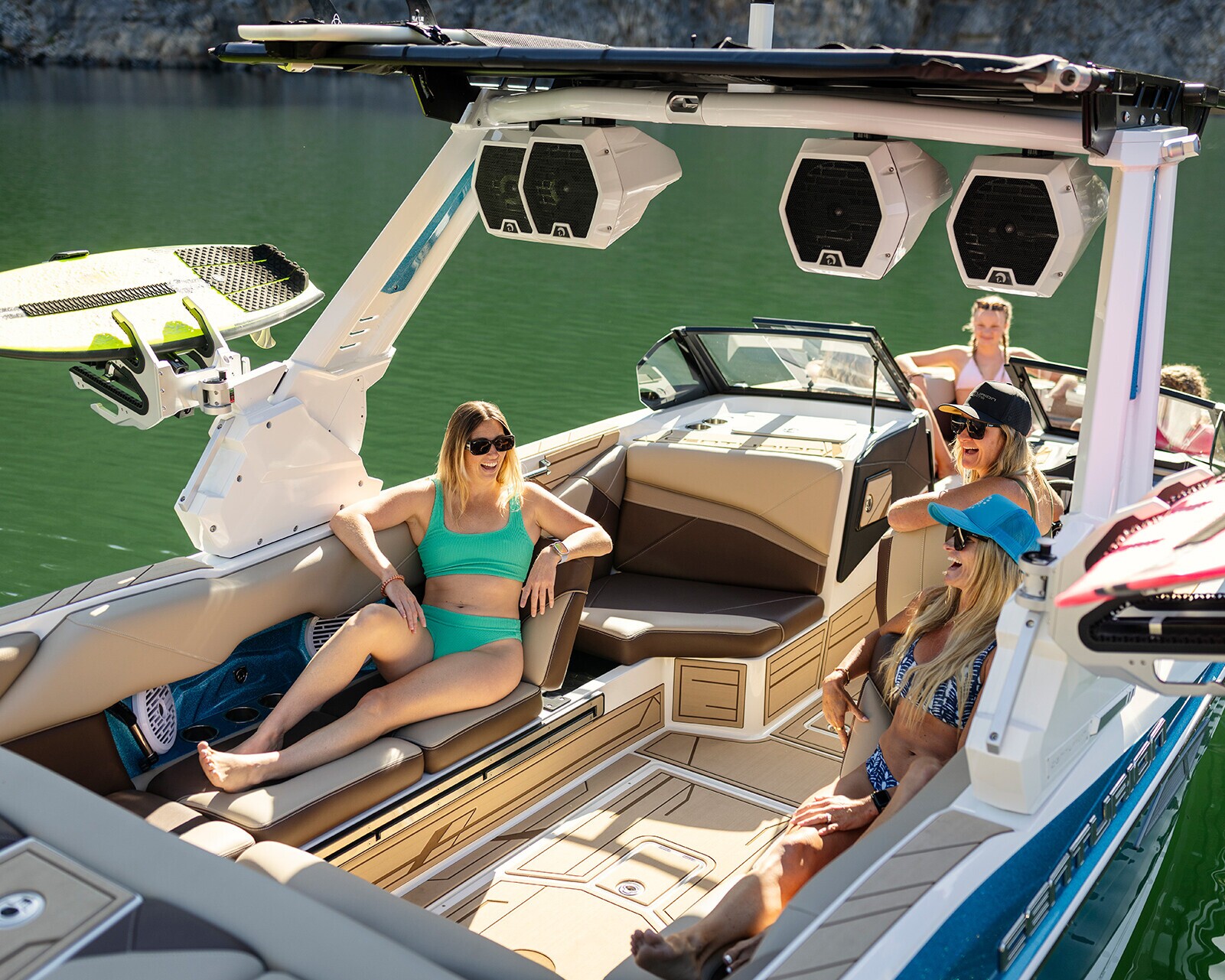 Four people relax and chat on a modern Centurion Nv243 Wake Boat in calm, green water, with wakeboards visible on board.