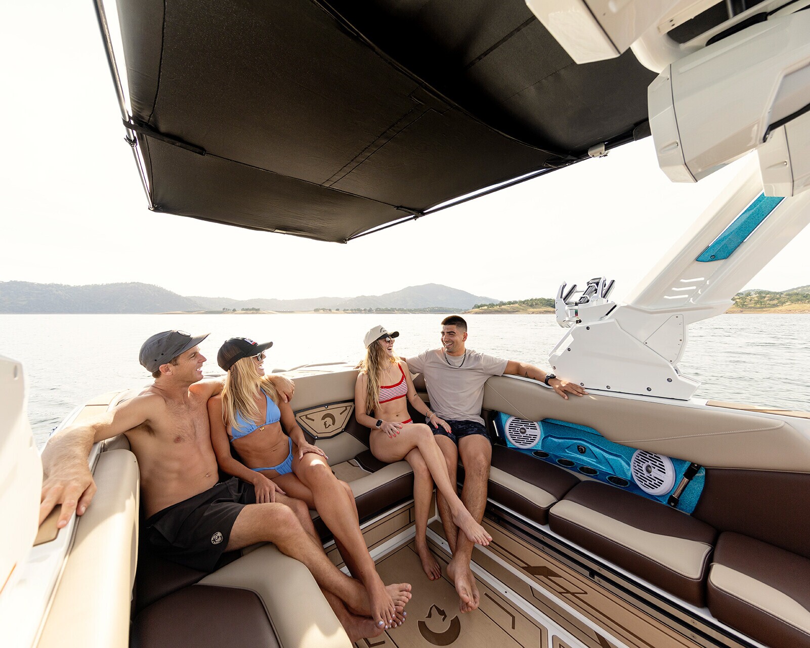 Four people in swimwear sit and talk on the seating area of a Centurion Nv243, with a lake and hills visible in the background.