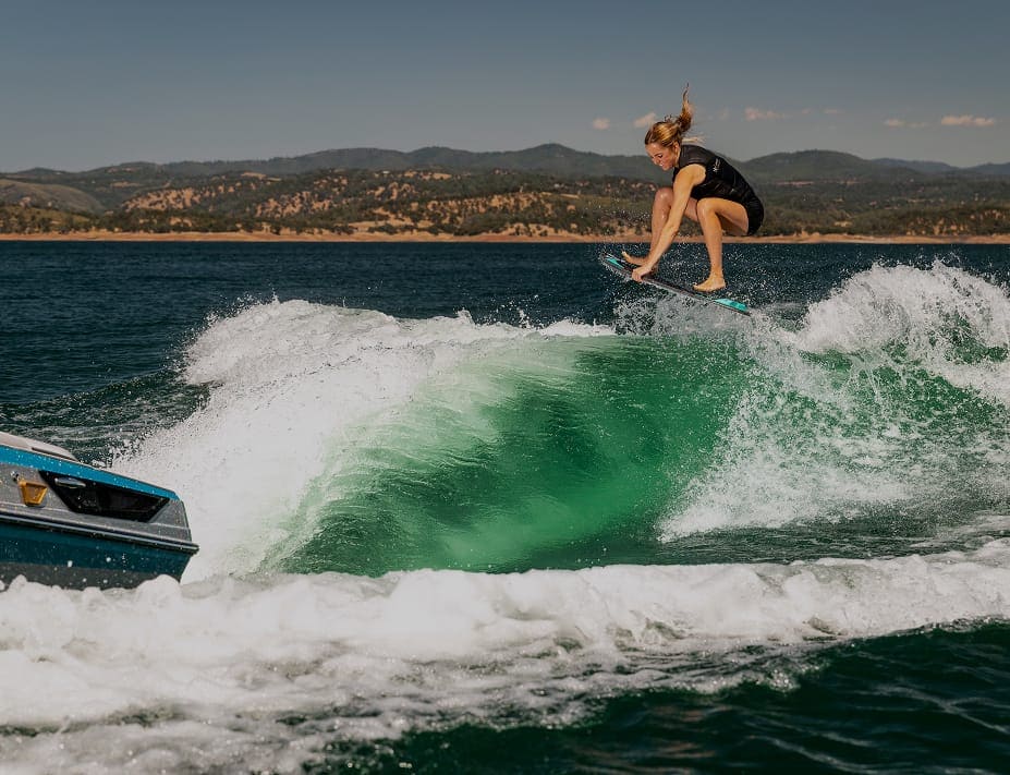 A person in a wetsuit is wakesurfing, catching air above the water behind a boat equipped with Centurion Ballast on a lake with hills in the background.