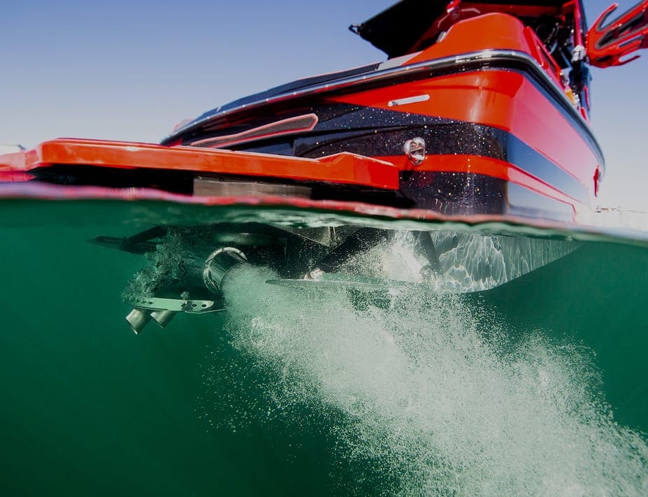 A red boat with a Centurion Opti-V Hull is partially submerged in water, bubbles trailing from its propeller, as seen from a split above-and-below water perspective.