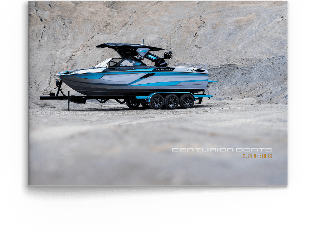 A blue and gray Centurion boat on a black trailer is parked on a rocky surface. Text reads &ldquo;Centurion Boats 2023 Ri Series&rdquo; in the lower right corner. Discover Centurion Boats&mdash;download the Centurion catalog for more details.