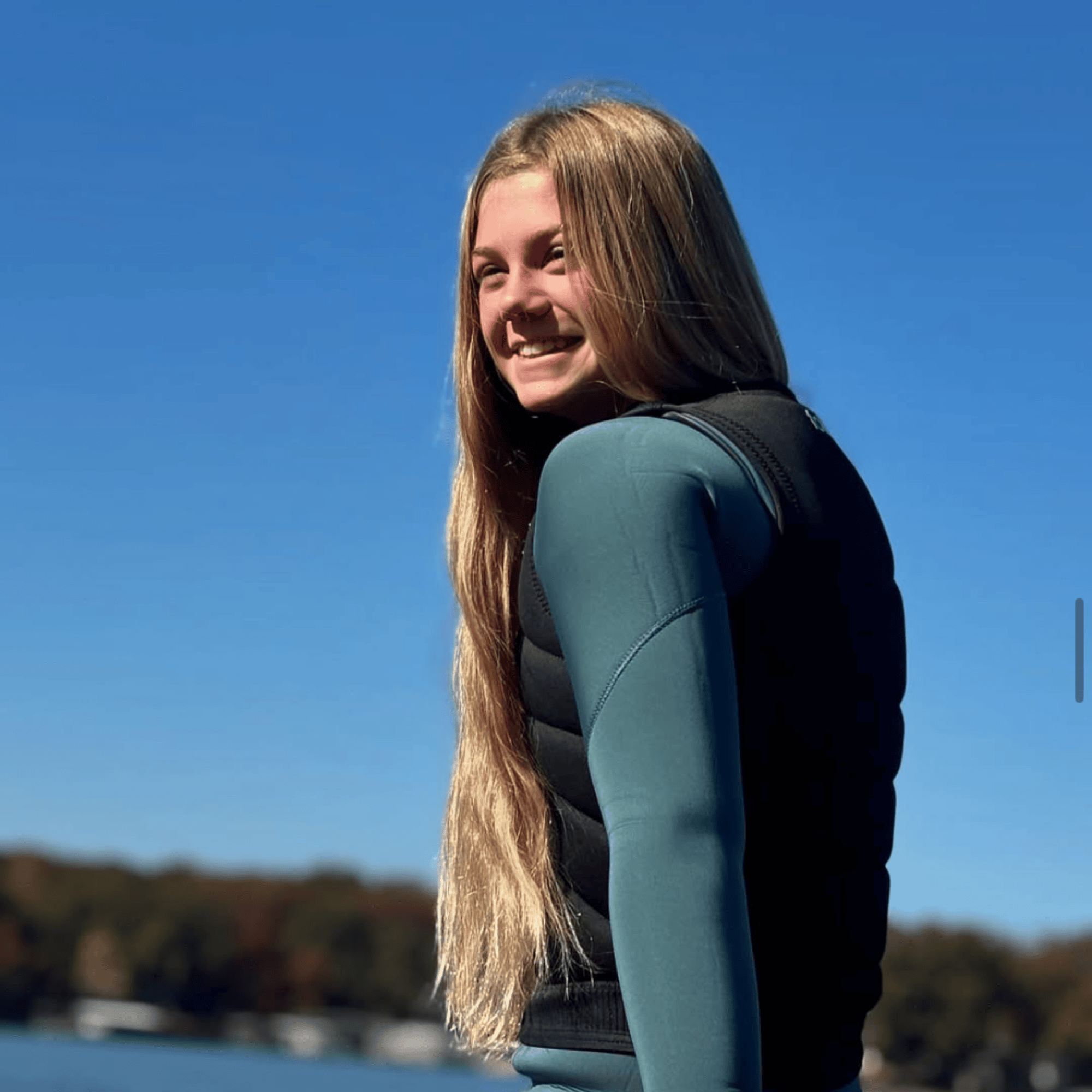 Dylan Ayala, a young person with long blonde hair wearing a wetsuit and life vest, stands by a body of water, smiling under a clear blue sky.