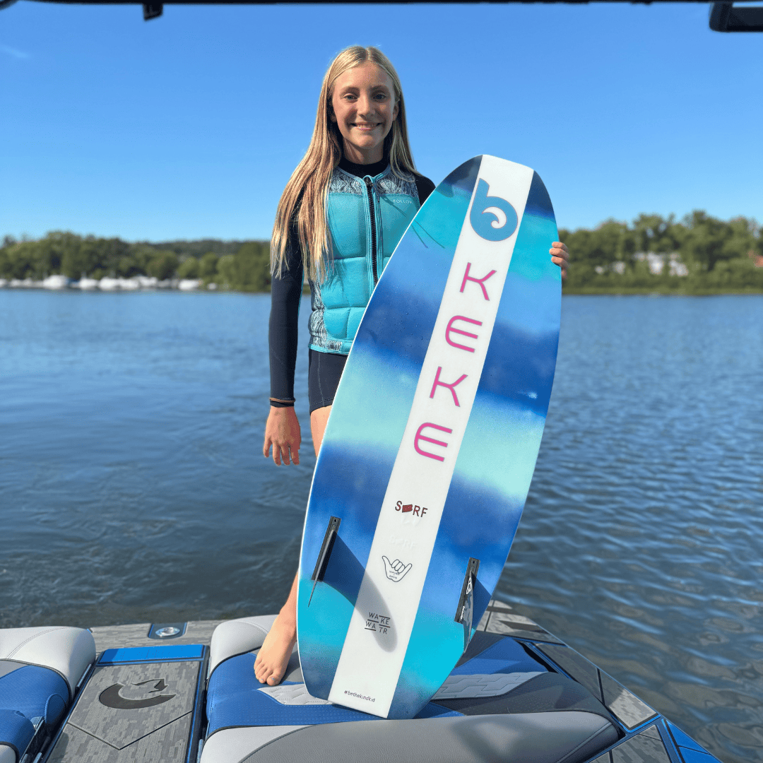 A girl in a blue life vest stands on a boat holding a blue and white surfboard with 