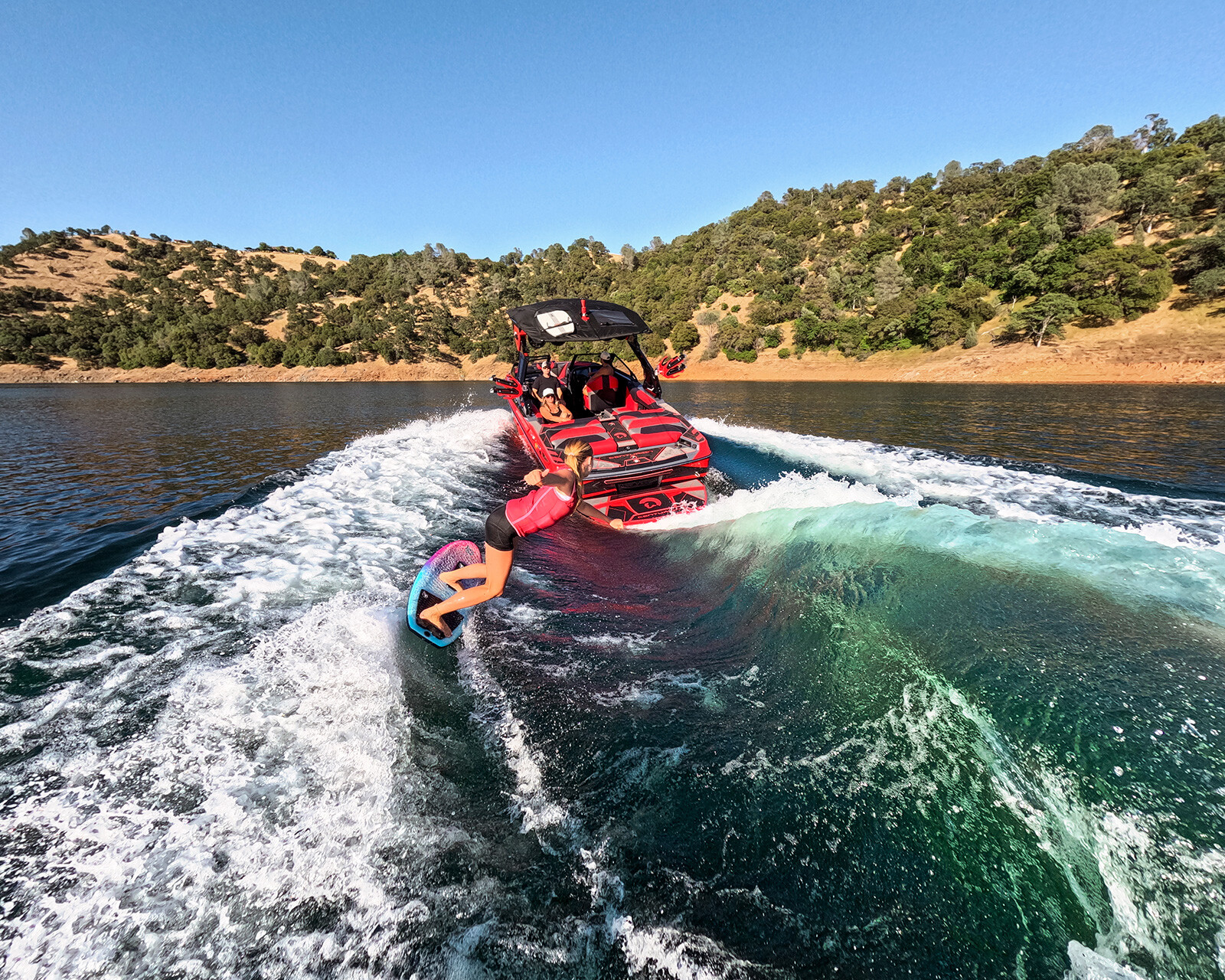 A person is wakesurfing behind a Centurion Ri230, a red surf boat, on a lake surrounded by tree-covered hills under a clear blue sky.