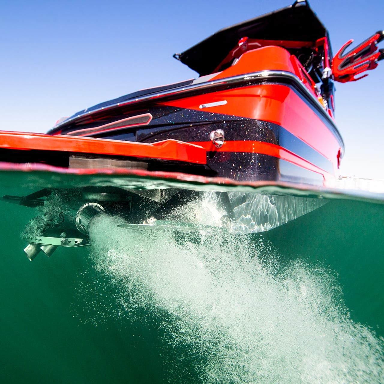 A red and black Centurion Ri230 Wake Boat is partially submerged in water, creating bubbles and turbulence behind its propeller, with a clear blue sky above.